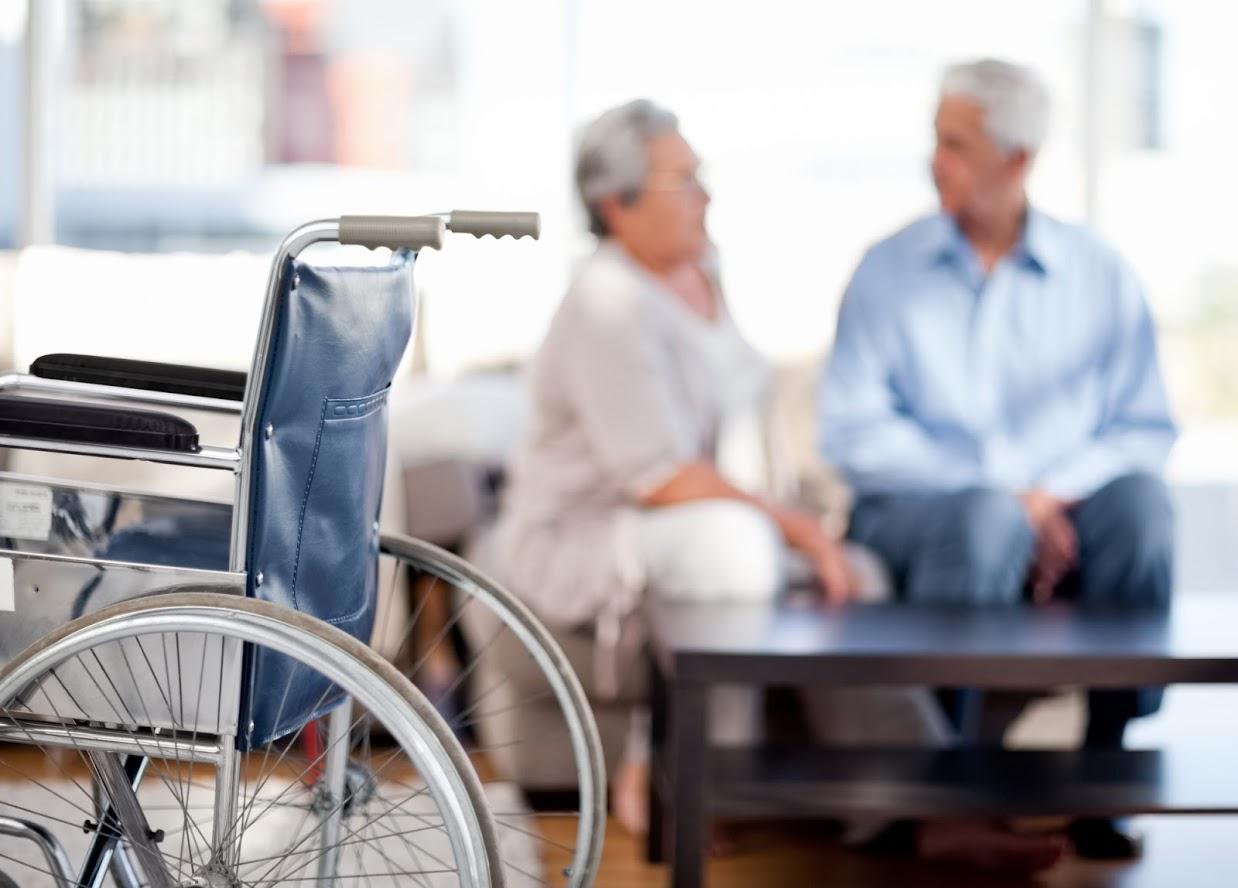 Wheelchair in foreground, blurred older couple seated on a sofa.