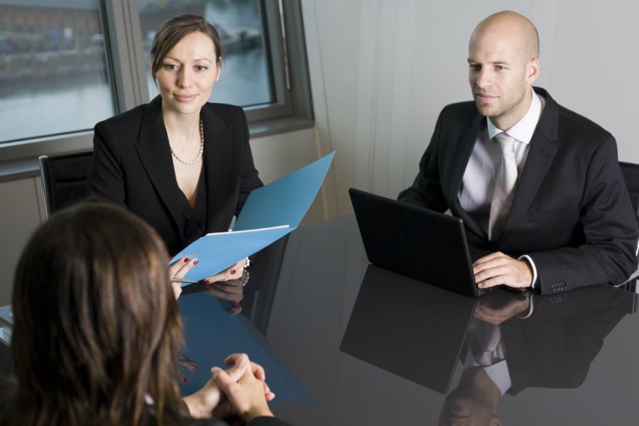 Two people interviewing a person at a table; one interviewer holds a file, the other uses a laptop.