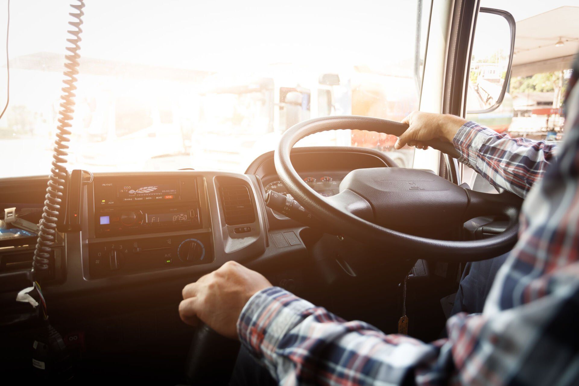 Driver in plaid shirt steering a vehicle, with bright sunlight through the windshield.