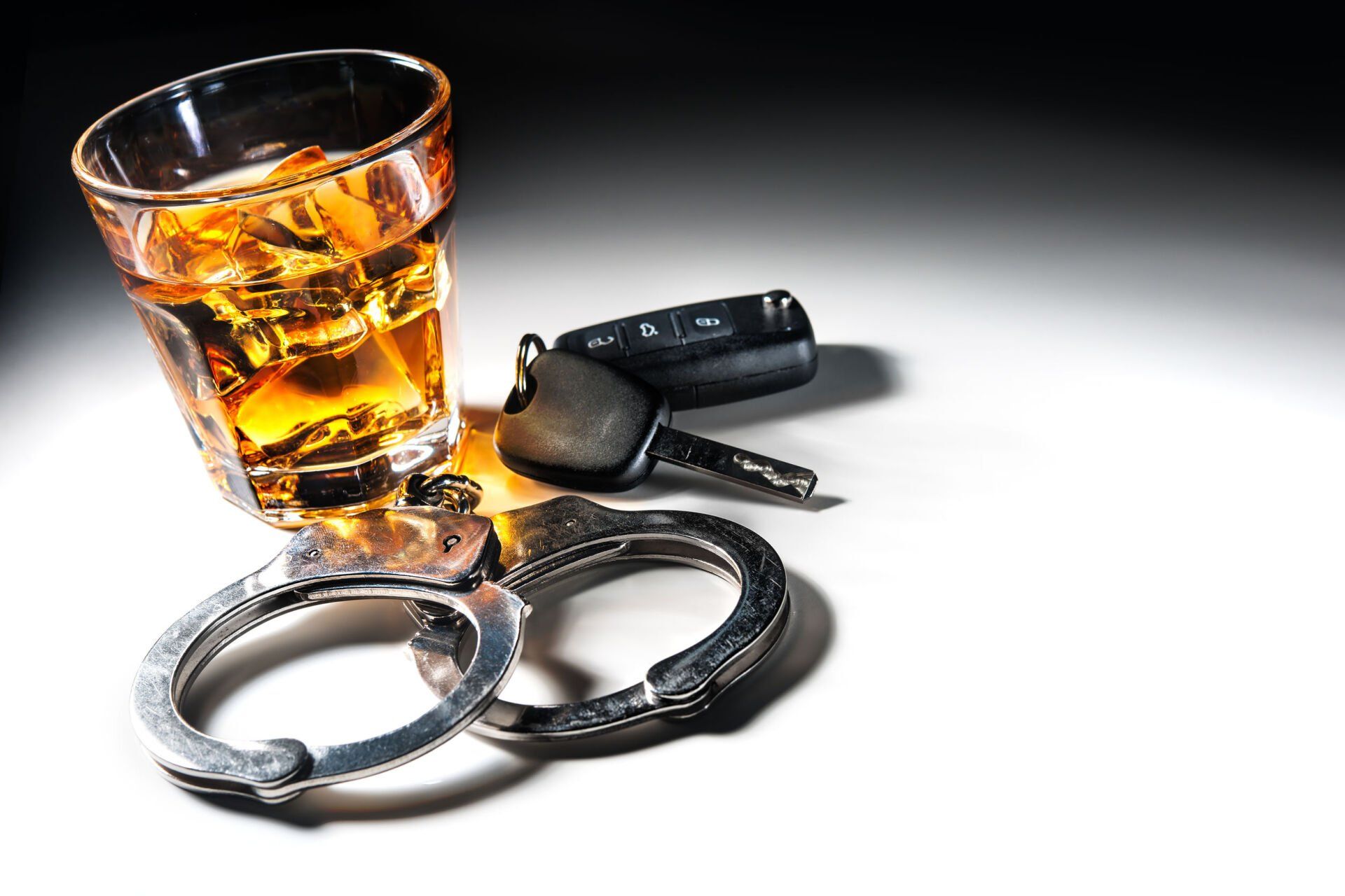 Glass of whiskey, car keys, and handcuffs on a white surface, suggesting drunk driving arrest