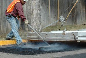Construction worker spreading asphalt on a road with a rake, orange vest, smoke, and blurred background.