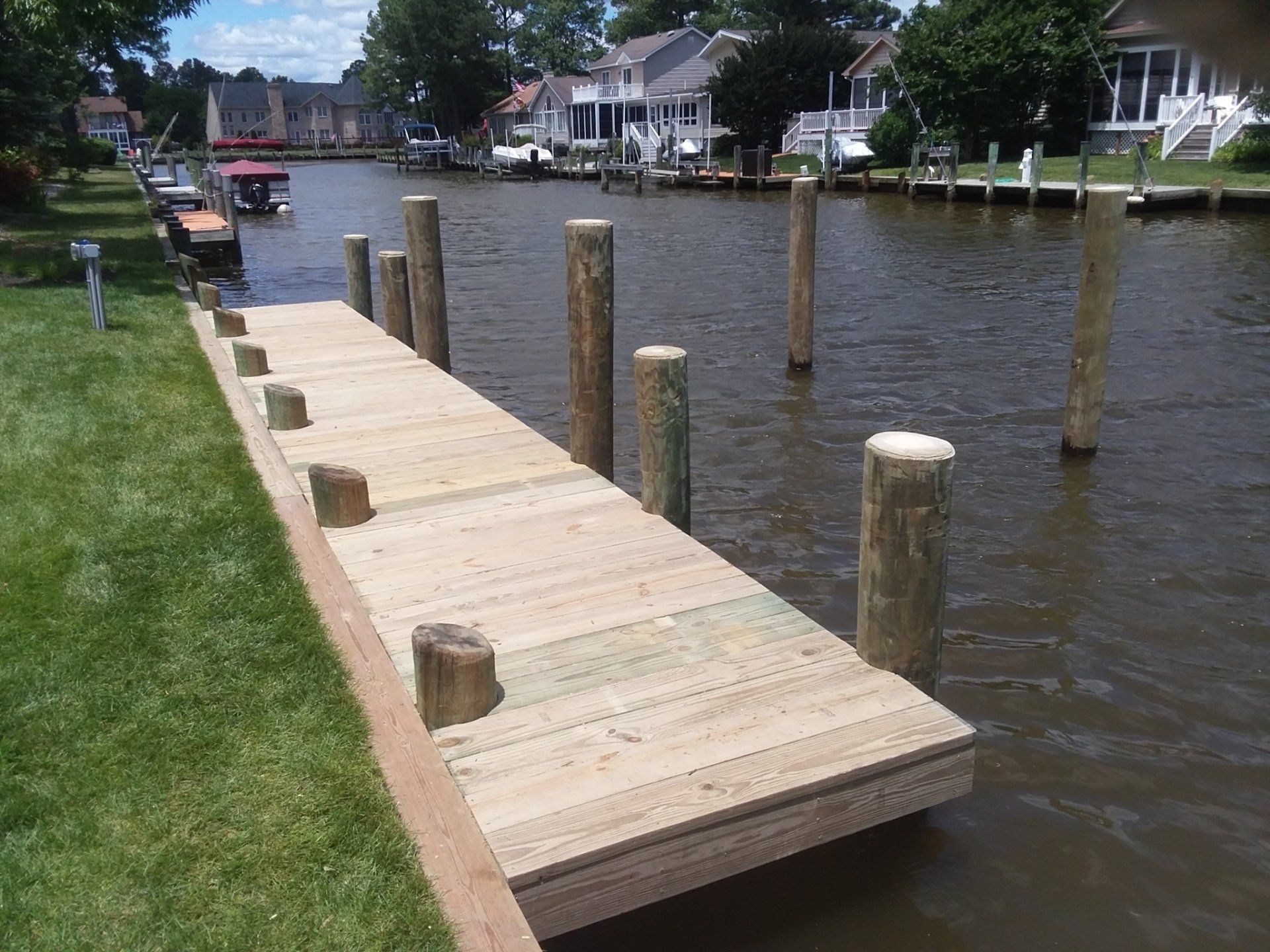 A newly constructed wooden dock extends into a narrow canal lined with houses and boat slips on a sunny day.