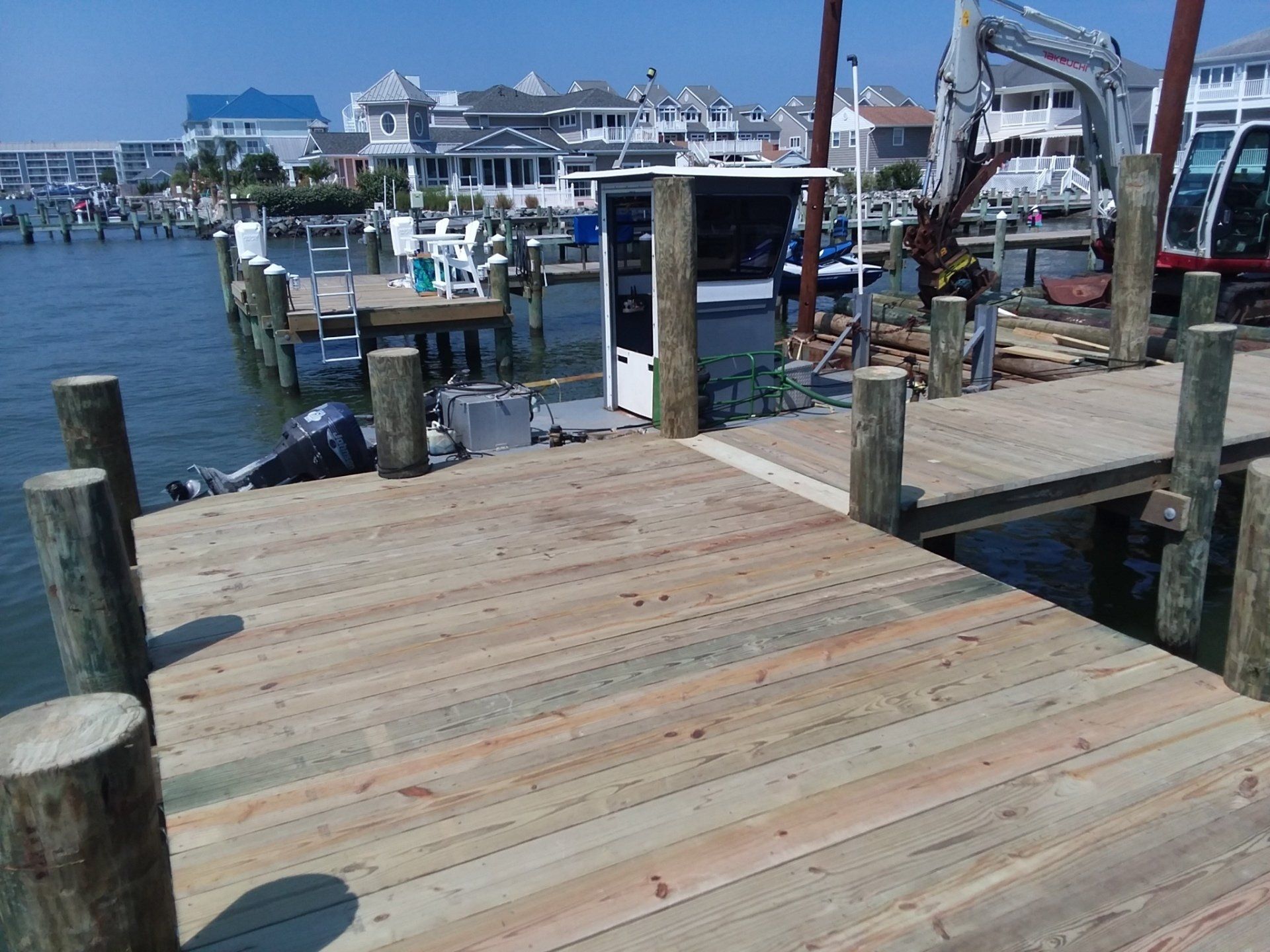 A wooden pier extends into a sunny bay with houses in the background and a construction machine parked near the docks.