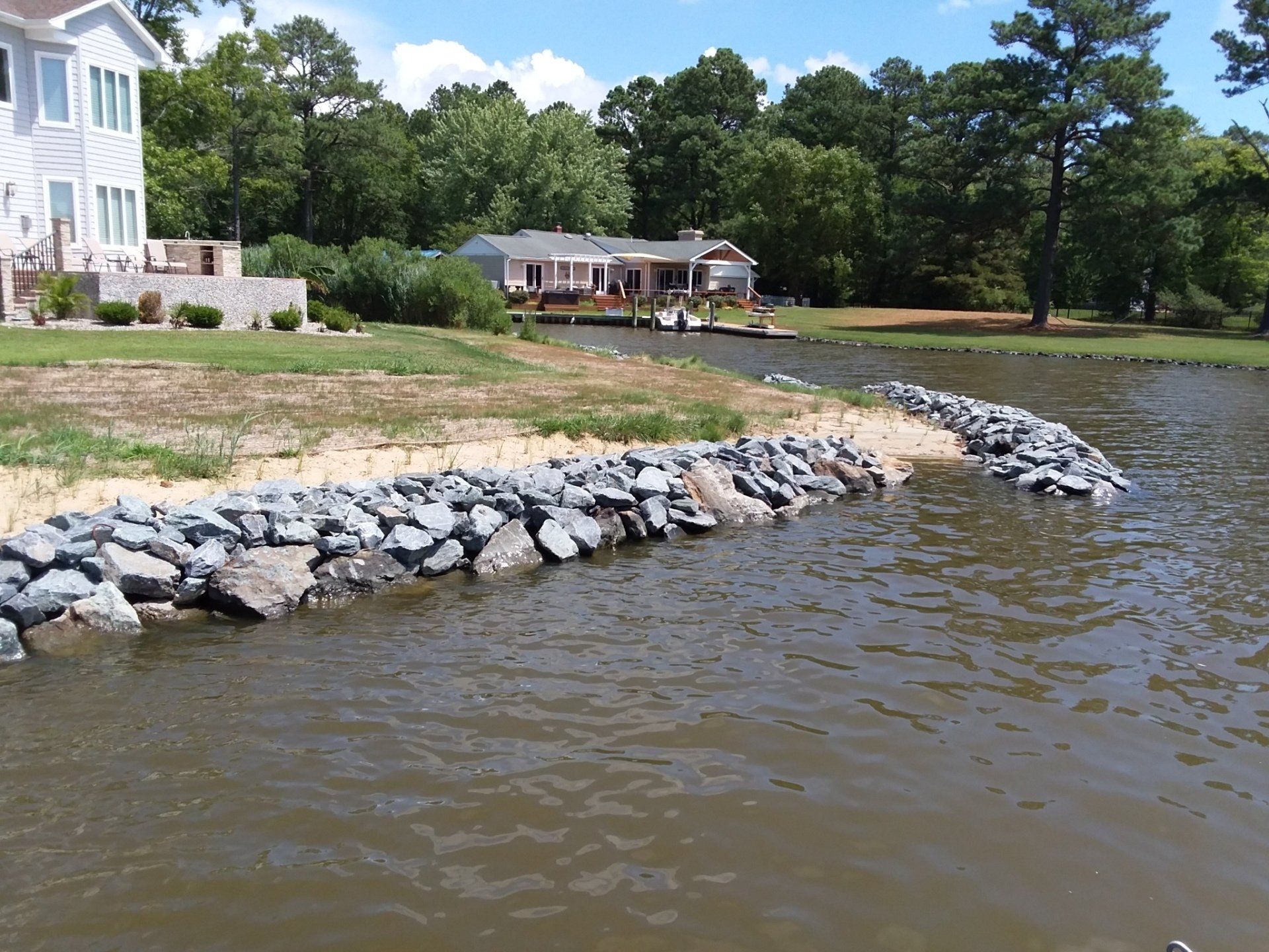 A shoreline featuring a gray stone riprap seawall protecting a sandy bank near a residential waterfront property.
