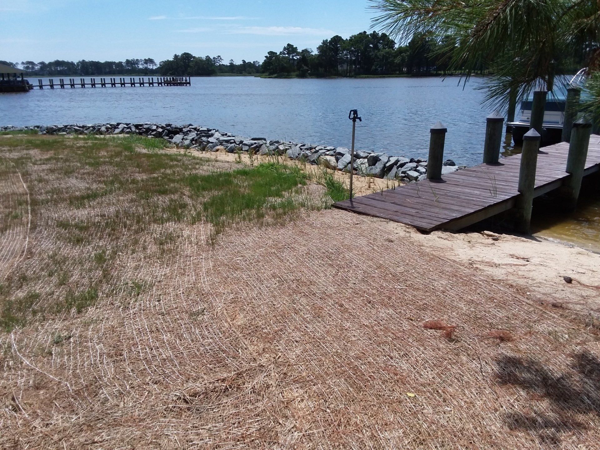A wooden dock extends into a calm lake from a shoreline covered in mulch and grass, with a rocky bulkhead along the water.