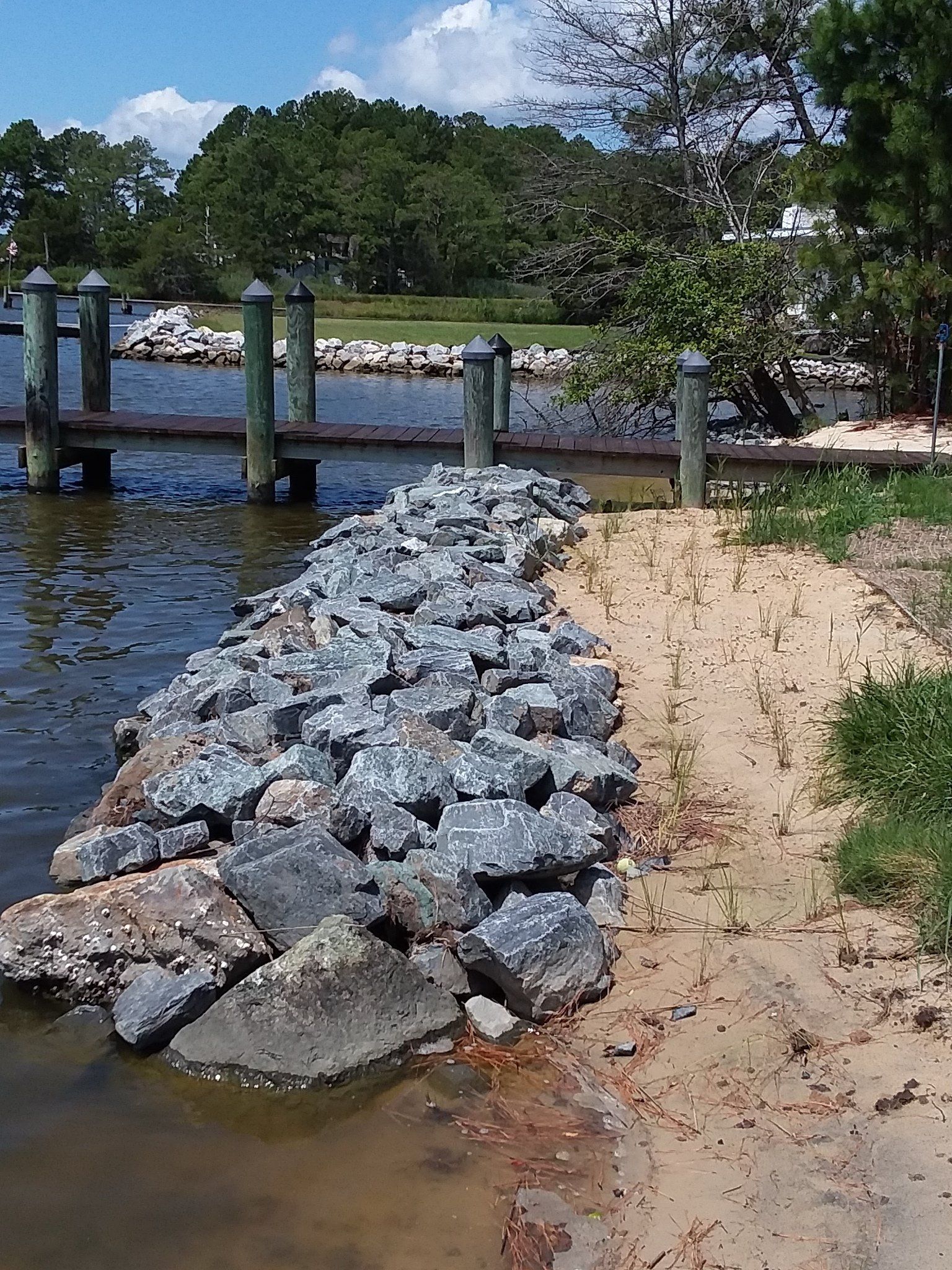 A pile of grey riprap rocks sits along a sandy shoreline near a wooden pier on a sunny day.