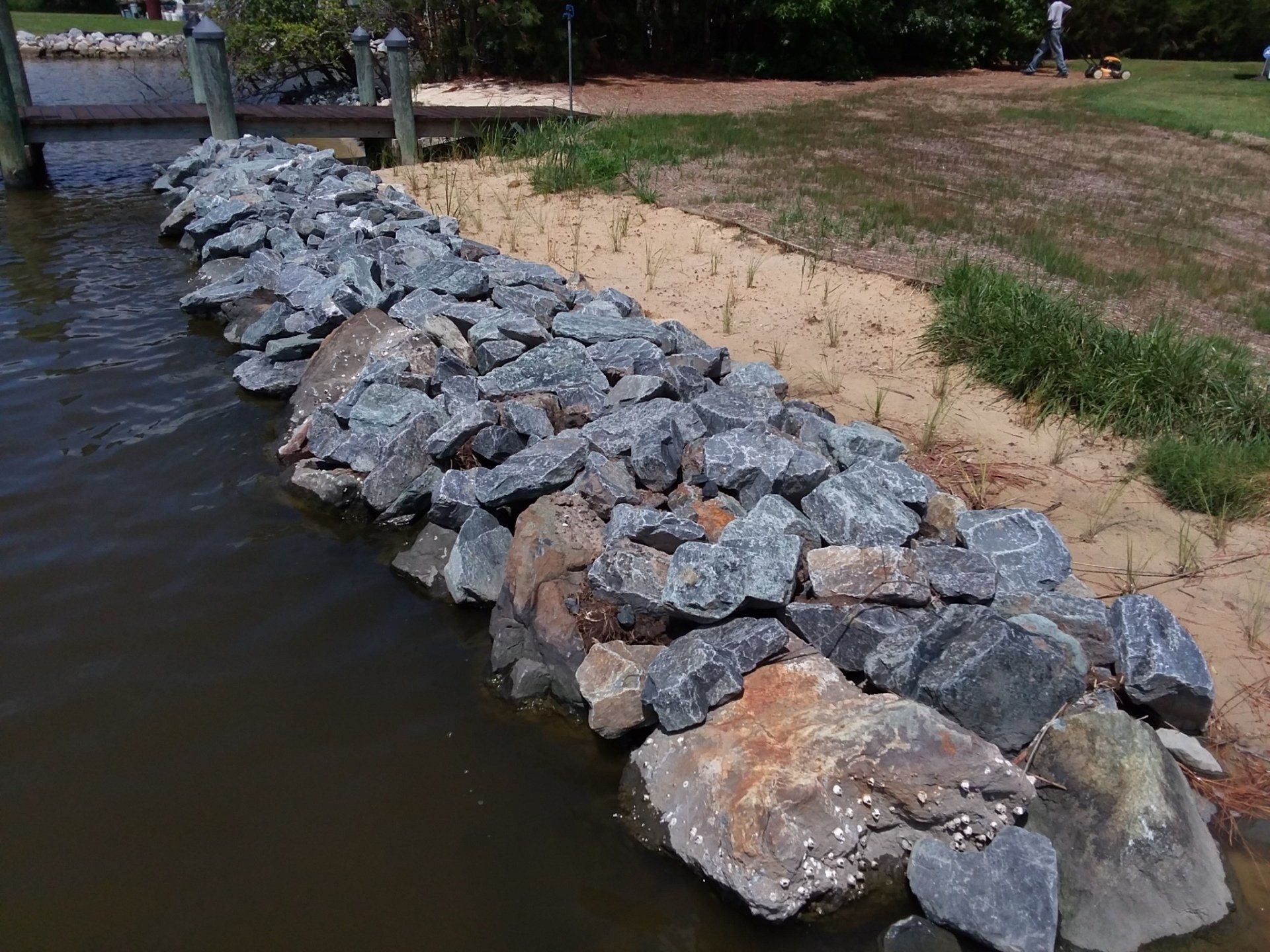 A riprap stone revetment protects a sandy shoreline from water erosion near a wooden pier.