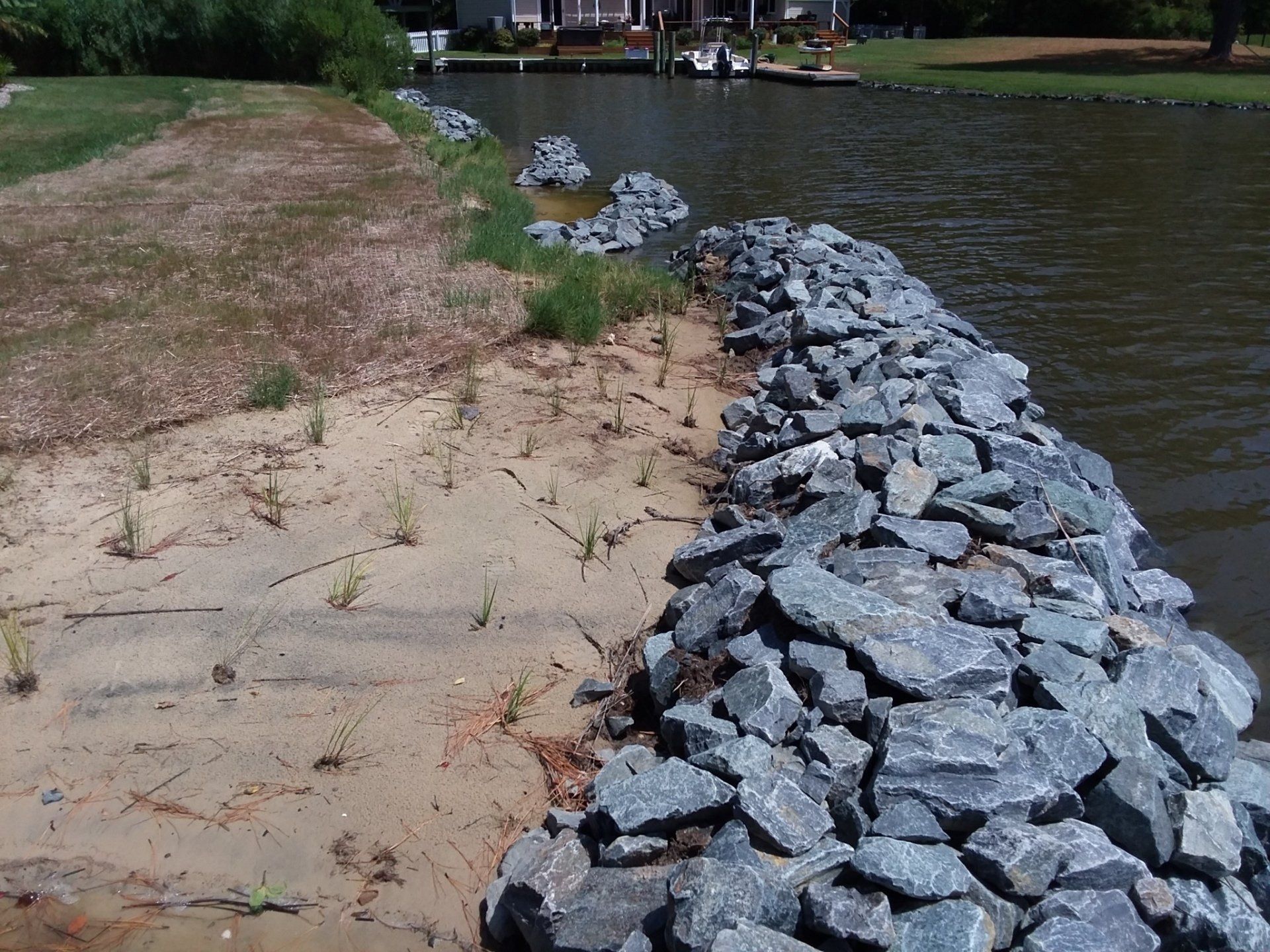 A riprap stone bulkhead protects a sandy shoreline along a waterway with houses in the background.