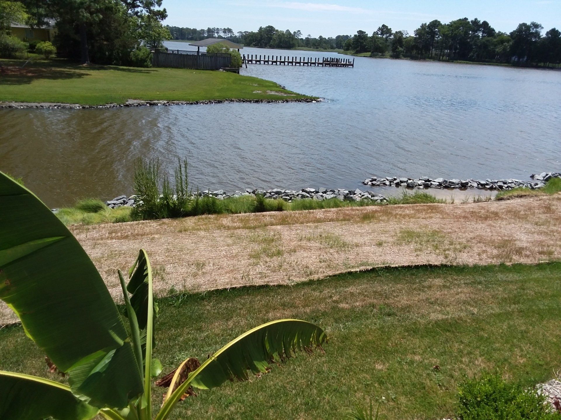 A green lawn slopes toward a calm body of water with a distant wooden dock under a clear blue sky.