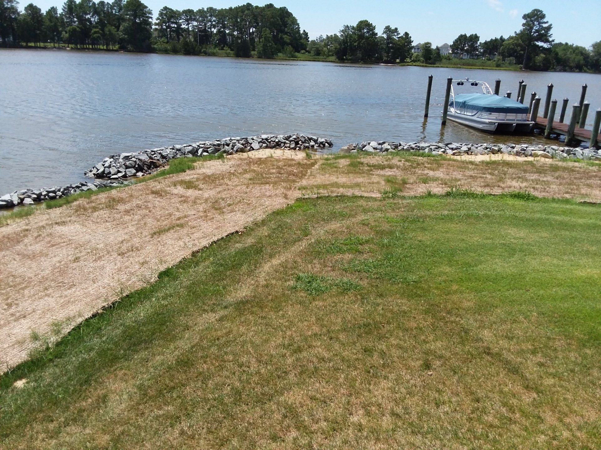 A shoreline with a gravel path and grassy lawn leading to a stone retaining wall at the edge of a calm, sunny lake.