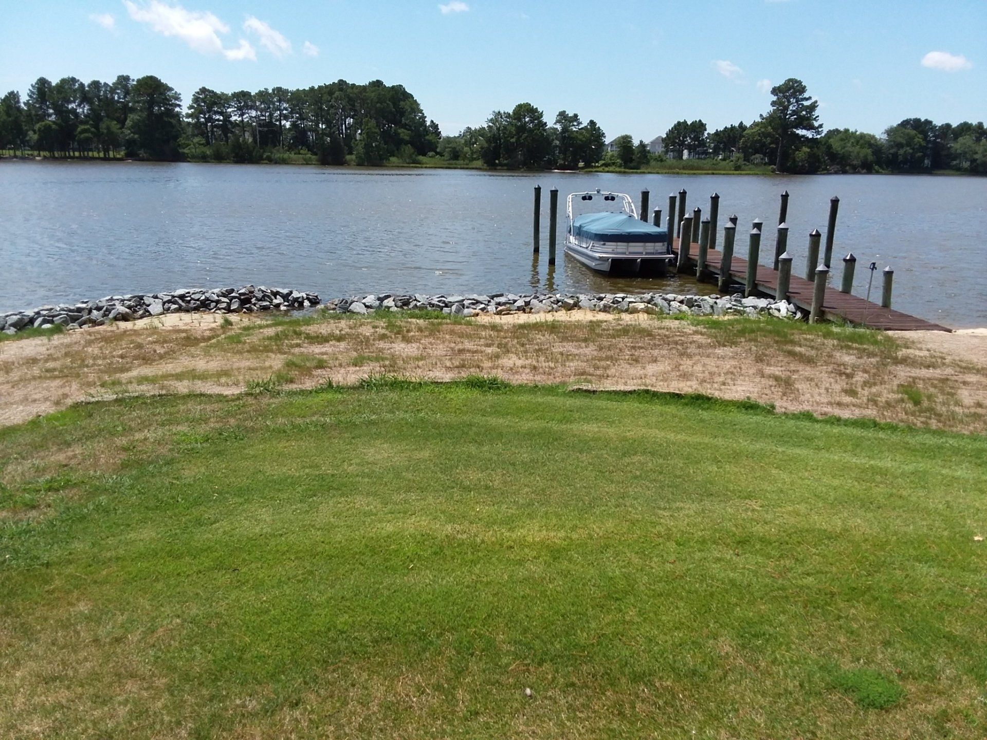 A pontoon boat moored at a wooden pier on a calm lake, with a green grassy lawn and rocky shoreline in the foreground.