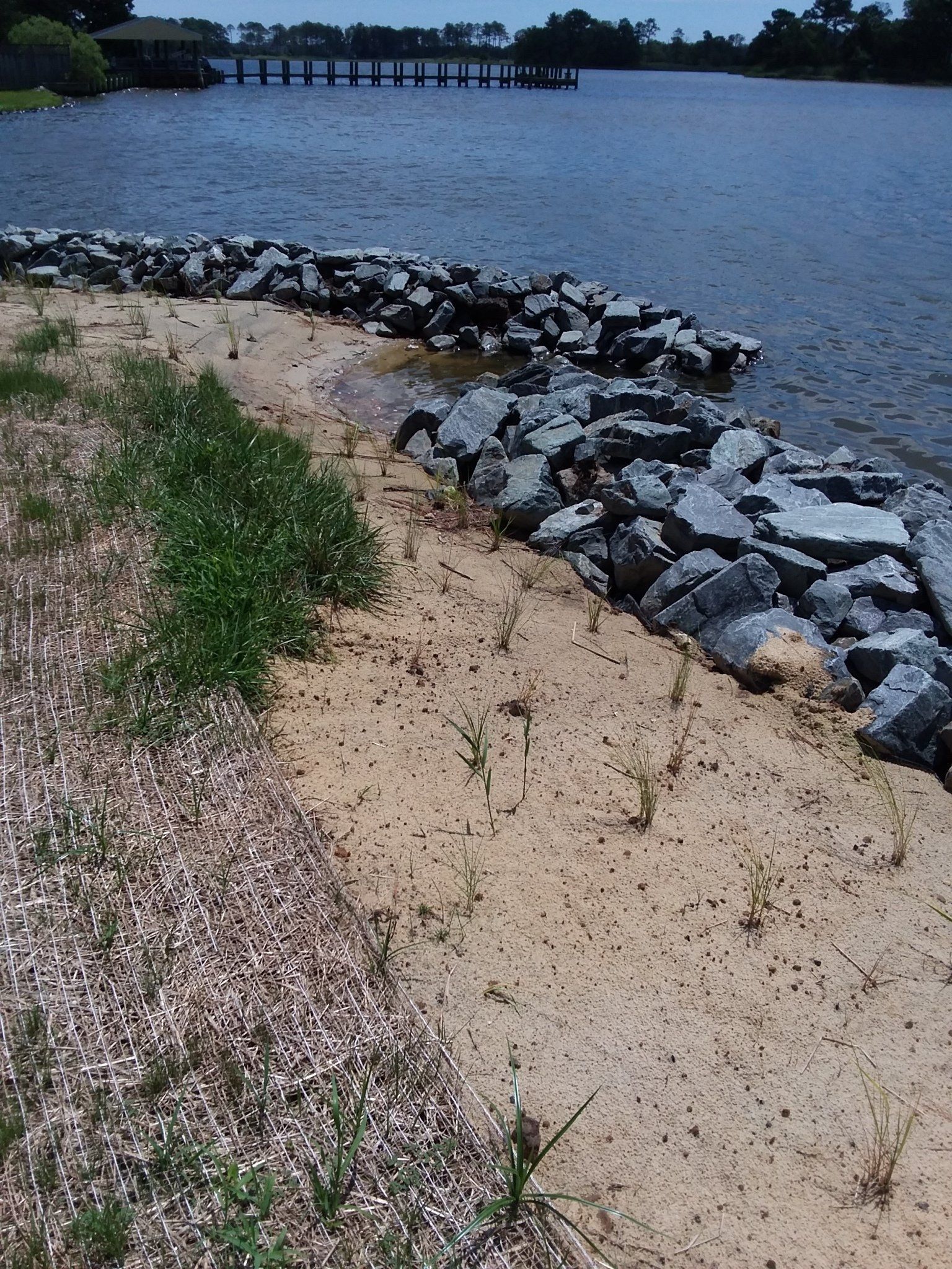 A sandy lakeshore lined with dark gray riprap rocks, featuring sparse green vegetation and a pier in the background.