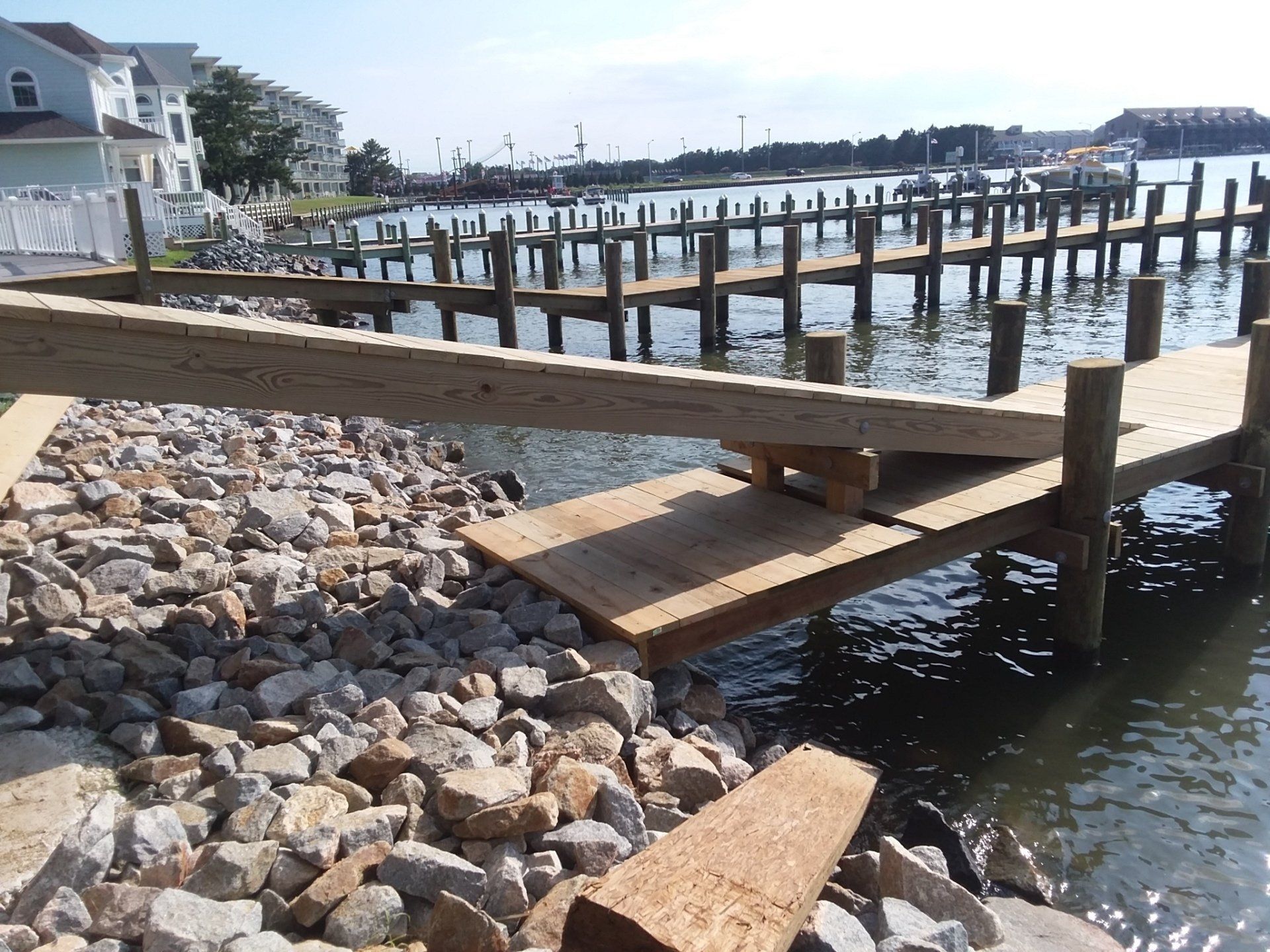 A damaged wooden pier with a large support beam detached and resting at an angle over a rocky shoreline and calm water.