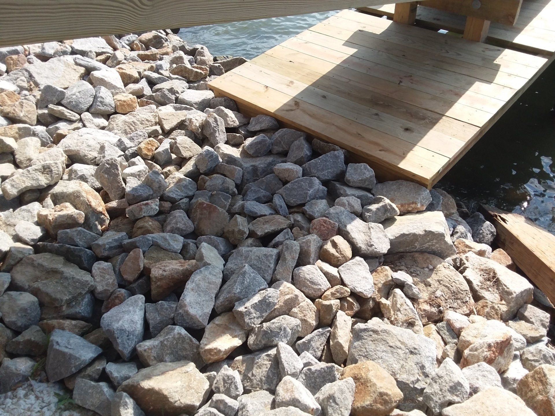 A pile of grey and tan rocks sits on a shoreline next to a wooden ramp leading into the water.