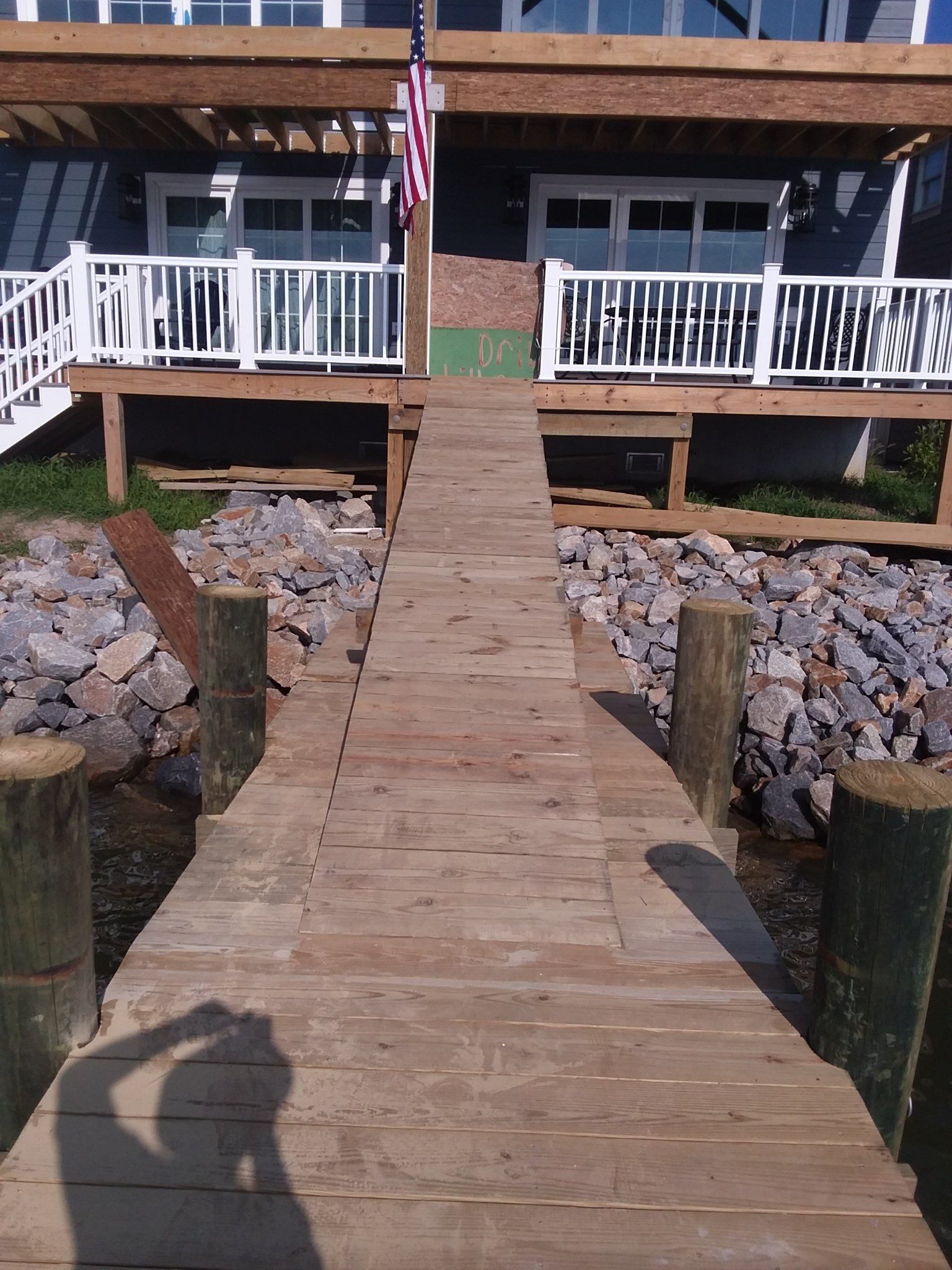 A wooden dock extends from the water toward a house with a raised deck and a small American flag.