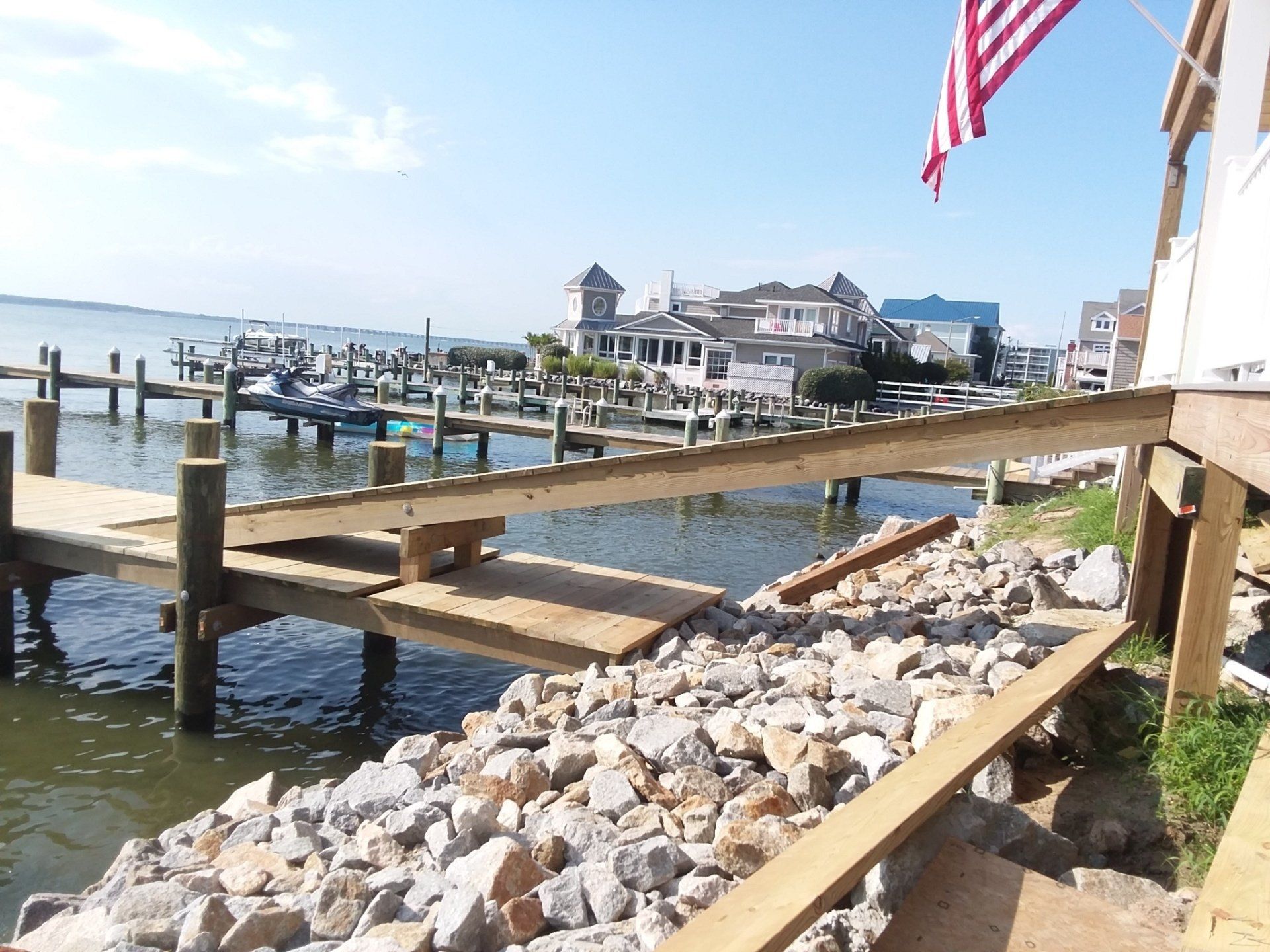 A wooden ramp leads from a rocky shoreline toward a wooden dock on a calm bay, with houses visible in the distance.