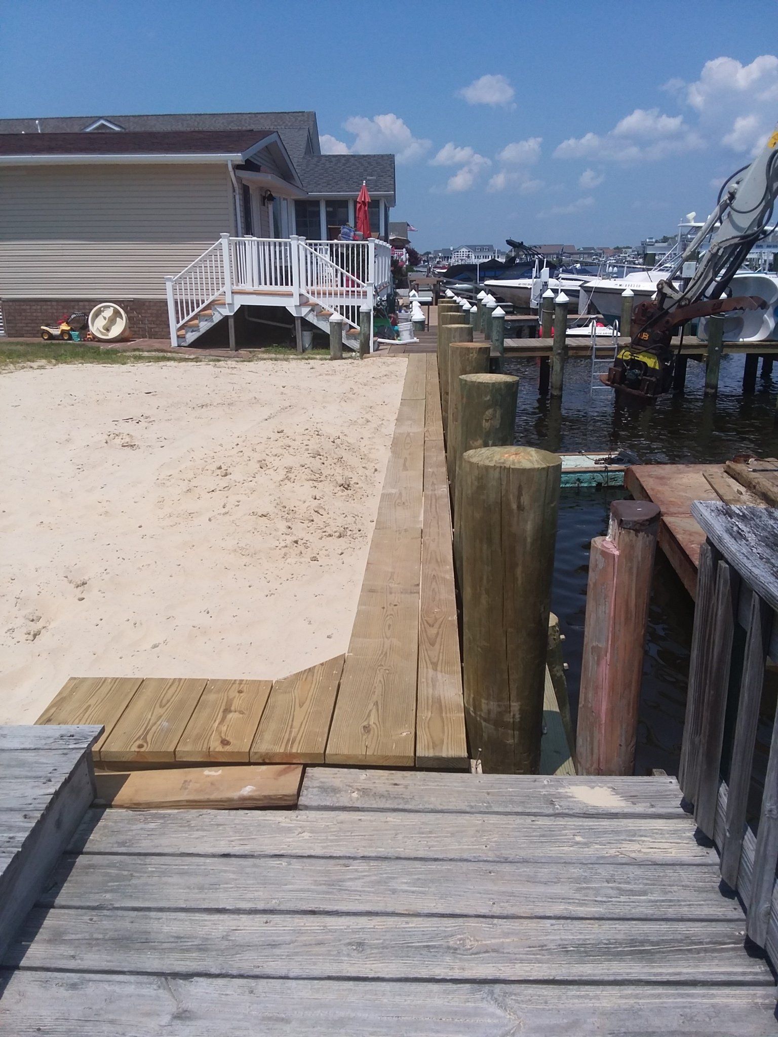 A wooden walkway runs along a sandy shoreline next to a wooden pier with pilings, with a house and boats in the background.