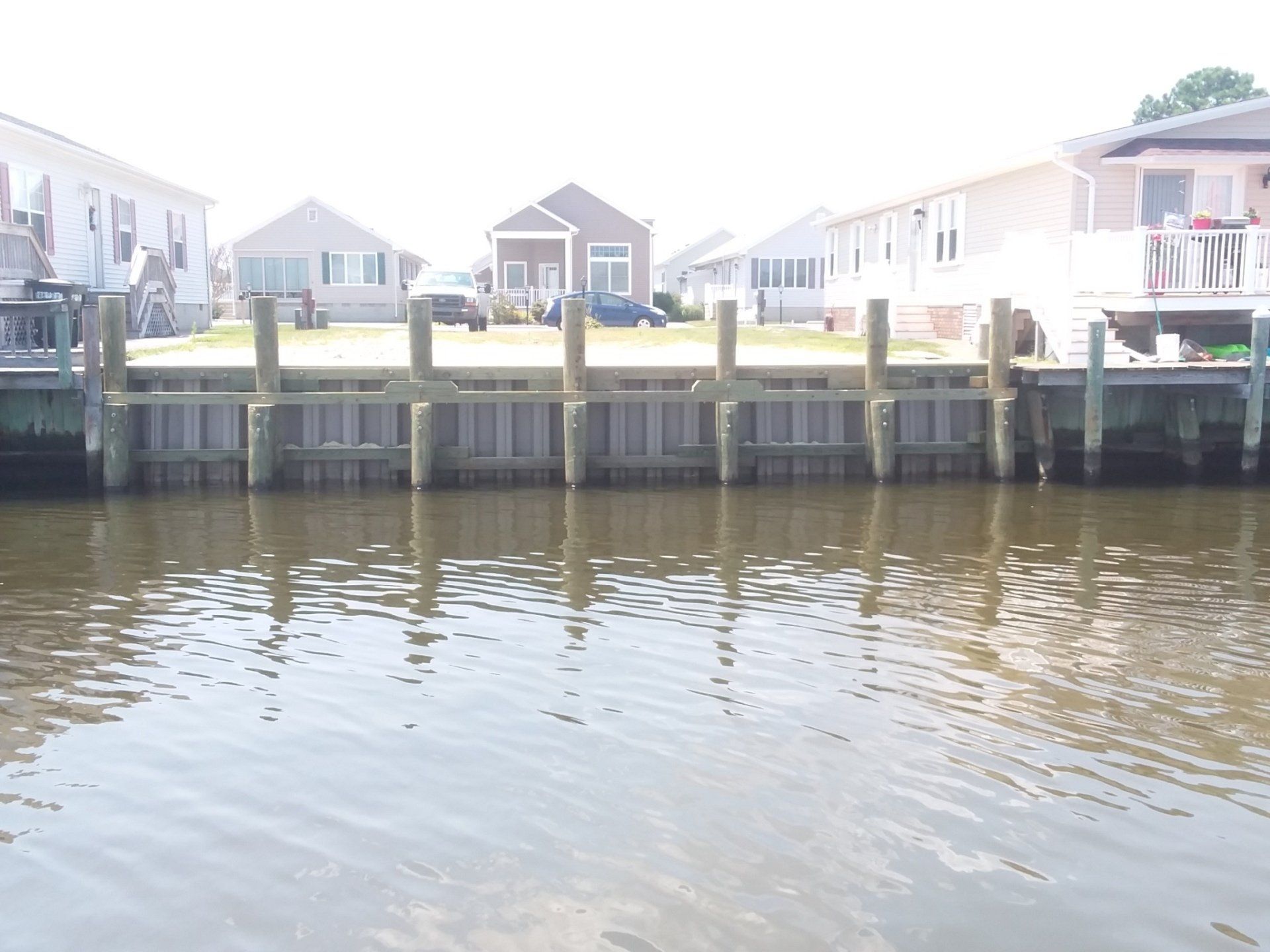 A bulkhead retaining wall made of vertical wooden posts and sheeting along a residential canal with houses in the back.