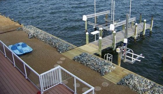 A baysidewooden dock and boat lift extend into blue water, adjacent to a gravel patio with a small blue pedal boat.