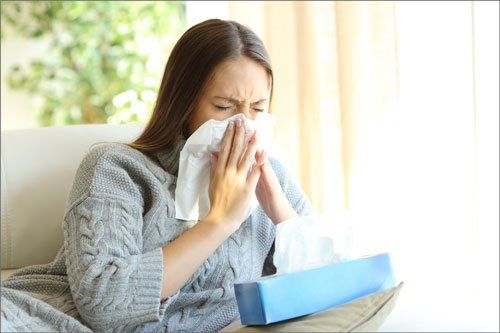 Woman in gray sweater sneezing into a tissue, with a box of tissues nearby.