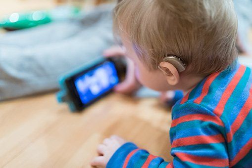 A toddler with a hearing aid watches a phone screen, indoors. They wear a blue and orange striped shirt.