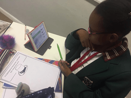 A young girl is sitting at a desk drawing a picture of a hat on a piece of paper.