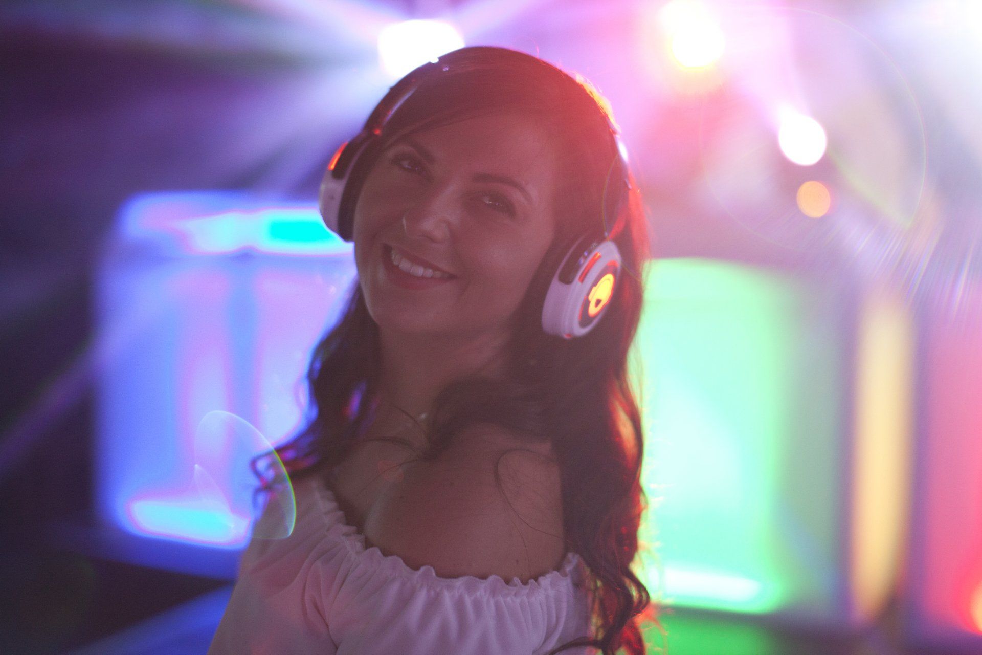 Woman with headphones smiles in a colorful, lit environment, possibly a club.