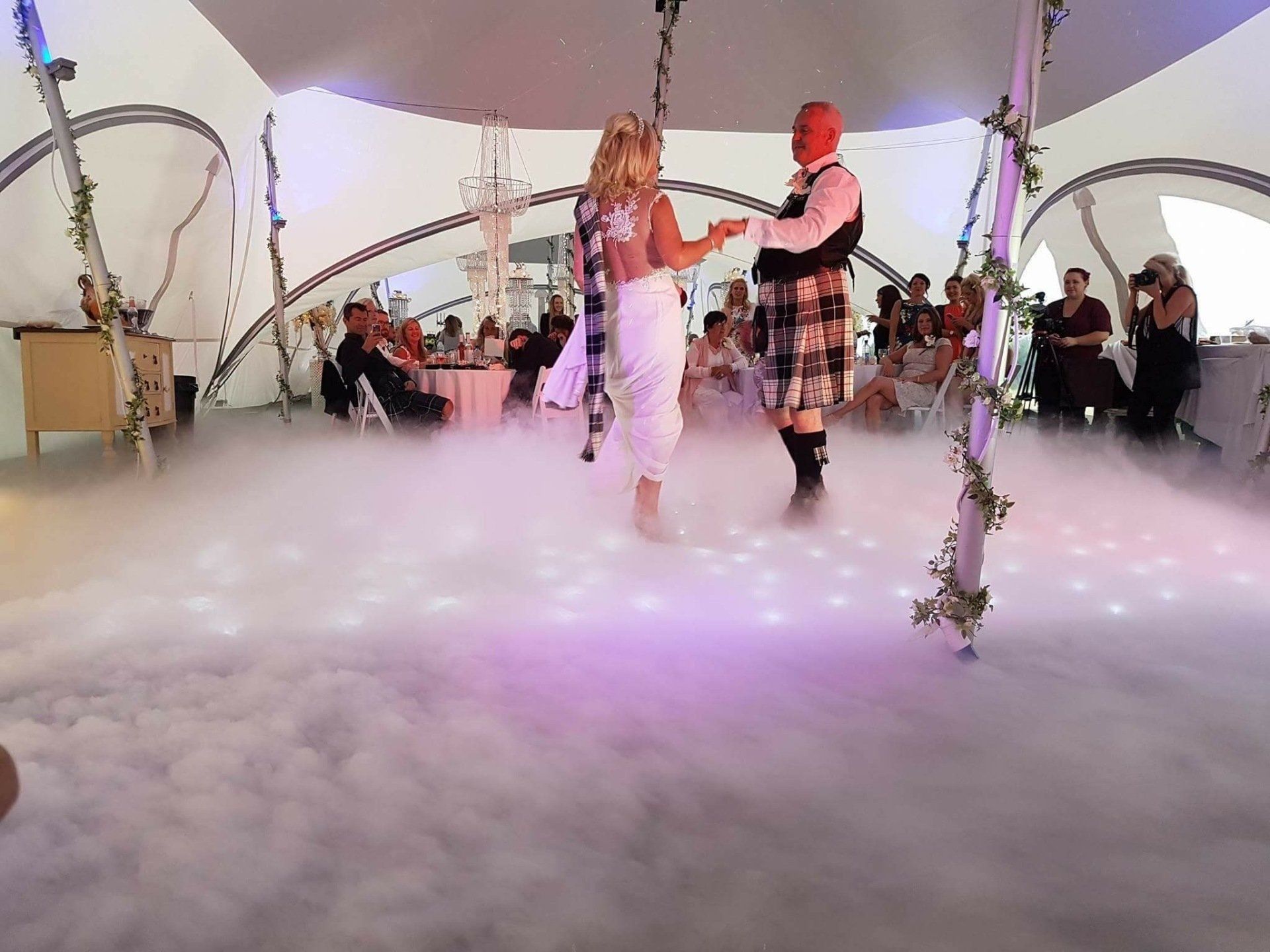 Bride and groom dance at a wedding reception, fog on the dance floor, guests seated at tables.