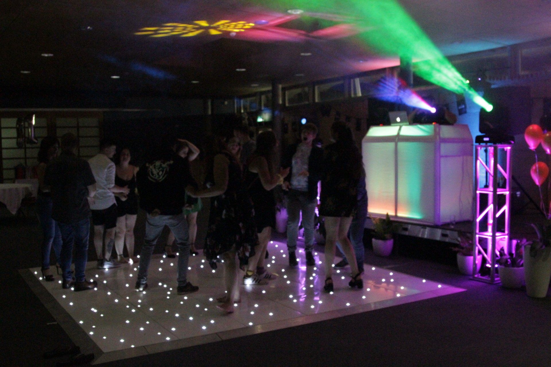 People dancing on a lit dance floor at a party, with colorful lights and a DJ booth.