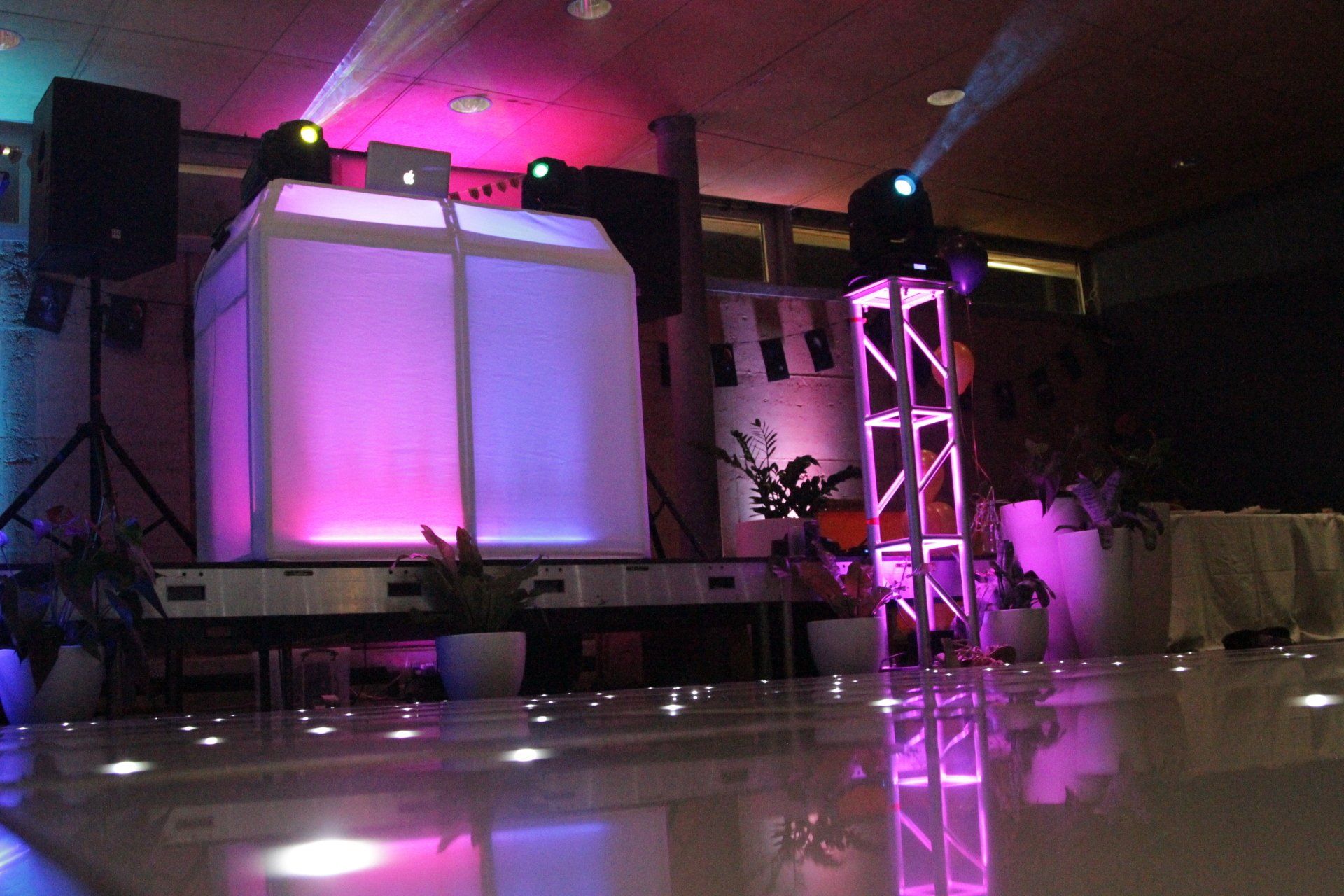 DJ setup at a party with light effects: white dance floor, illuminated cube DJ booth, speakers, and stage lights.