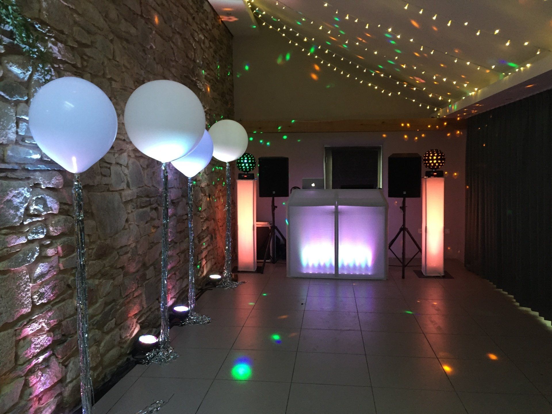 DJ setup with white balloons, colorful lights, and speakers in a party room.