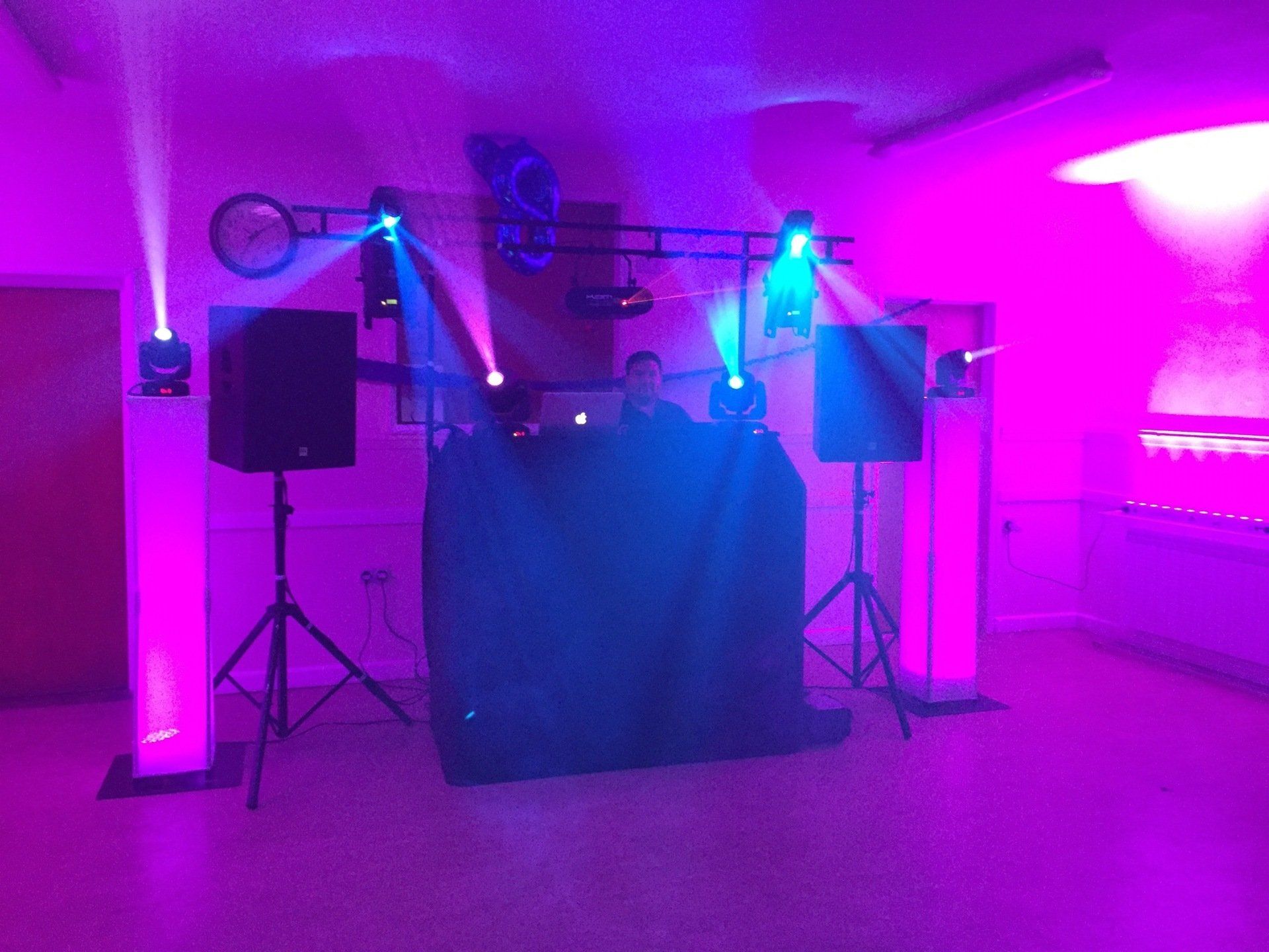 A DJ setup in a room lit with purple and blue lights.  The DJ is behind a table.  Speakers are on stands.