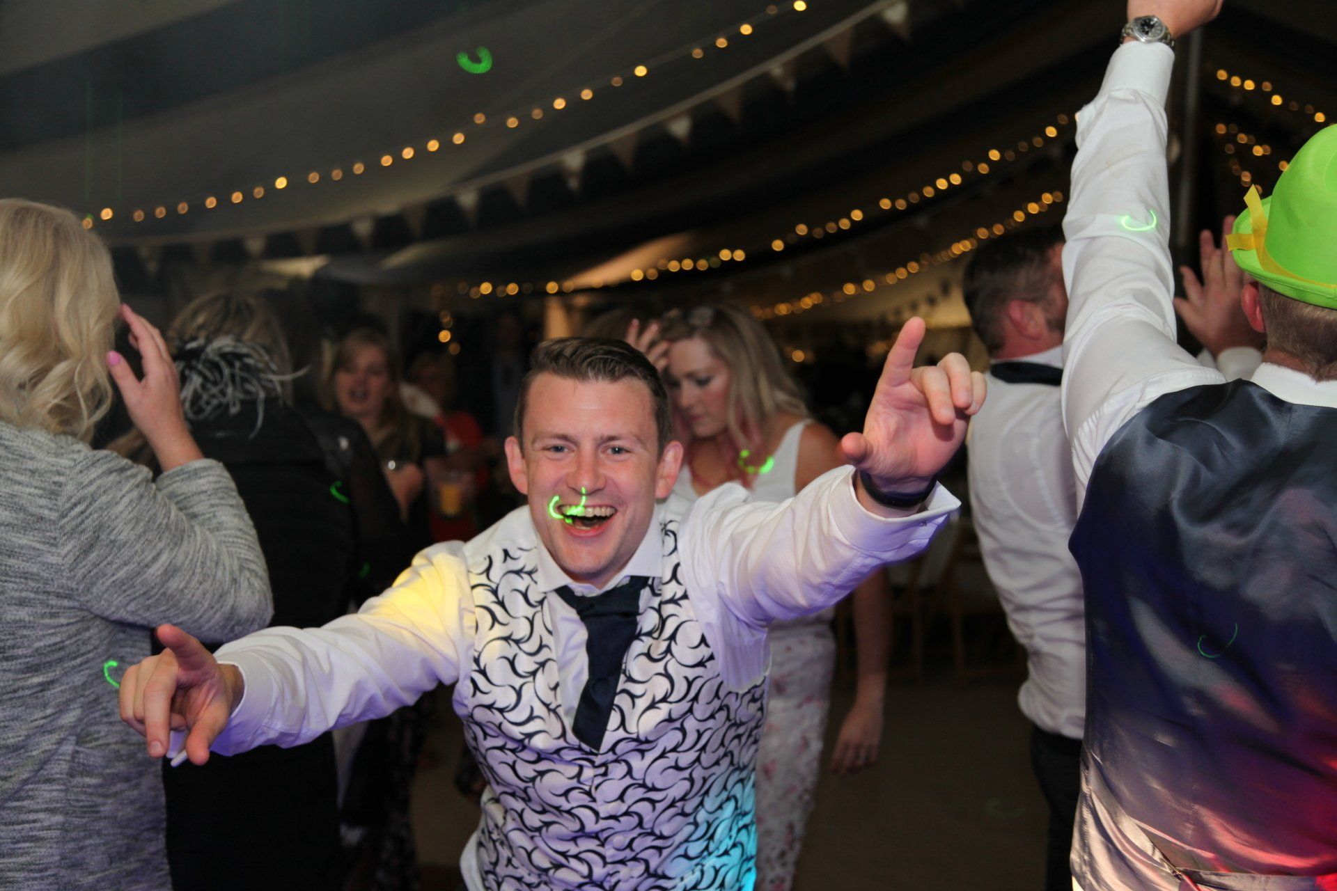 Man dancing, arms raised, wearing a white vest and tie, in a crowded party.
