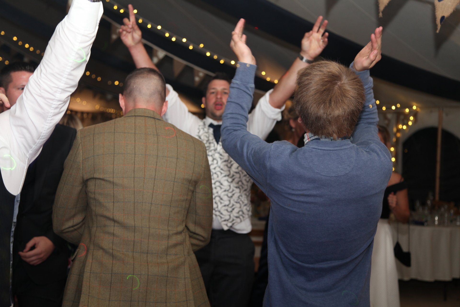 People dancing at a celebration, arms raised. Men in suits inside a tent with string lights.