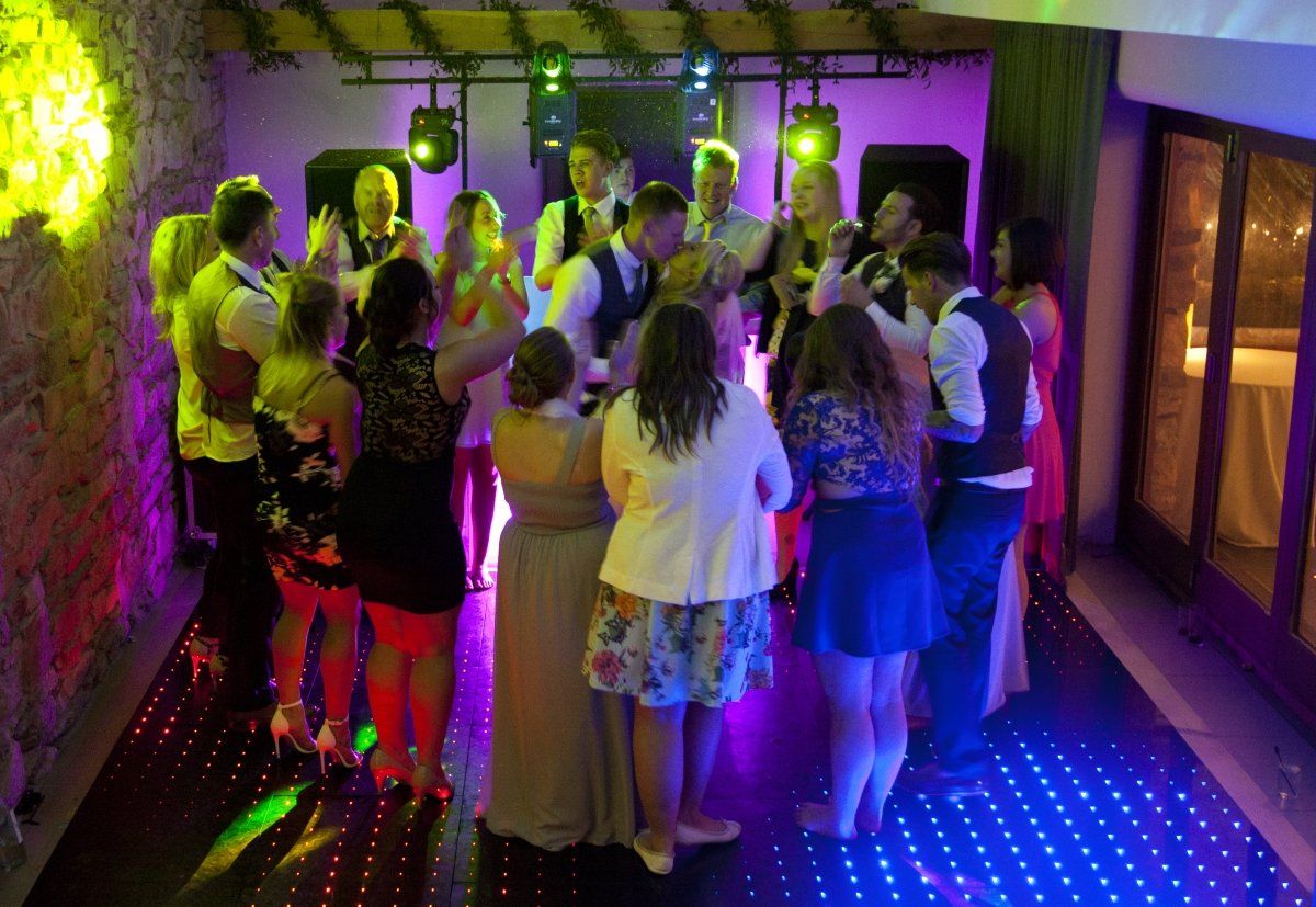 People dancing at a party; a band plays on a small stage with stage lights; a wood-paneled window.