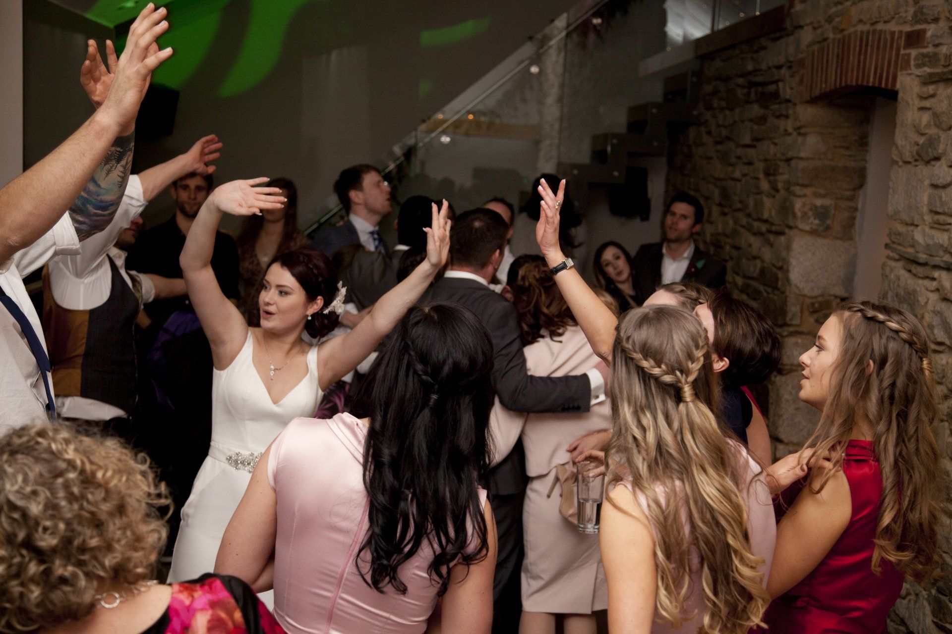 Wedding reception with dancing people. Bride with arms up, others with raised arms, brick wall in background.