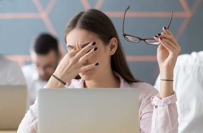 Frustrated worker holding glasses behind a laptop in a blurred office