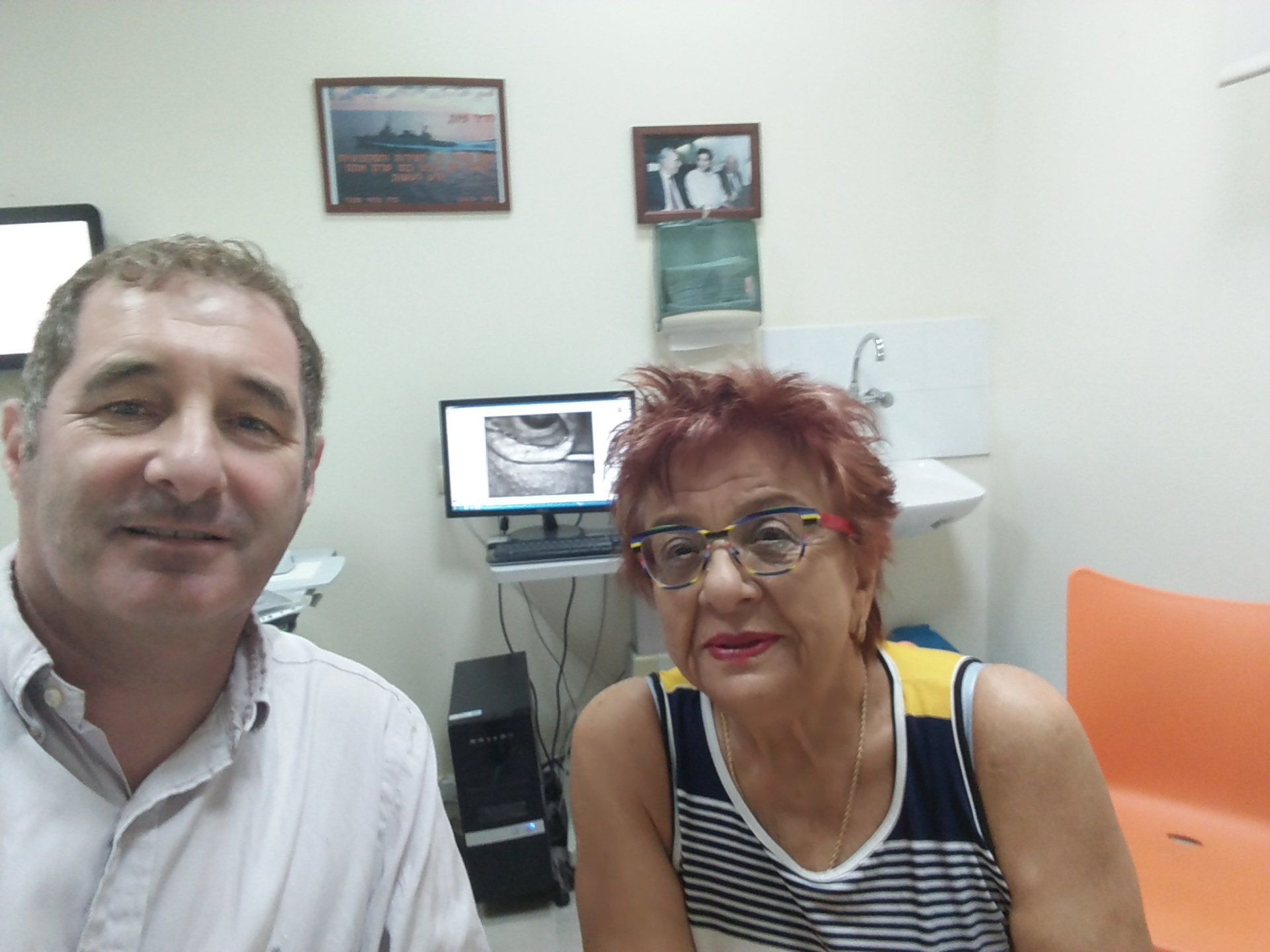 Two people smiling in an office, with a computer and orange chair in the background.
