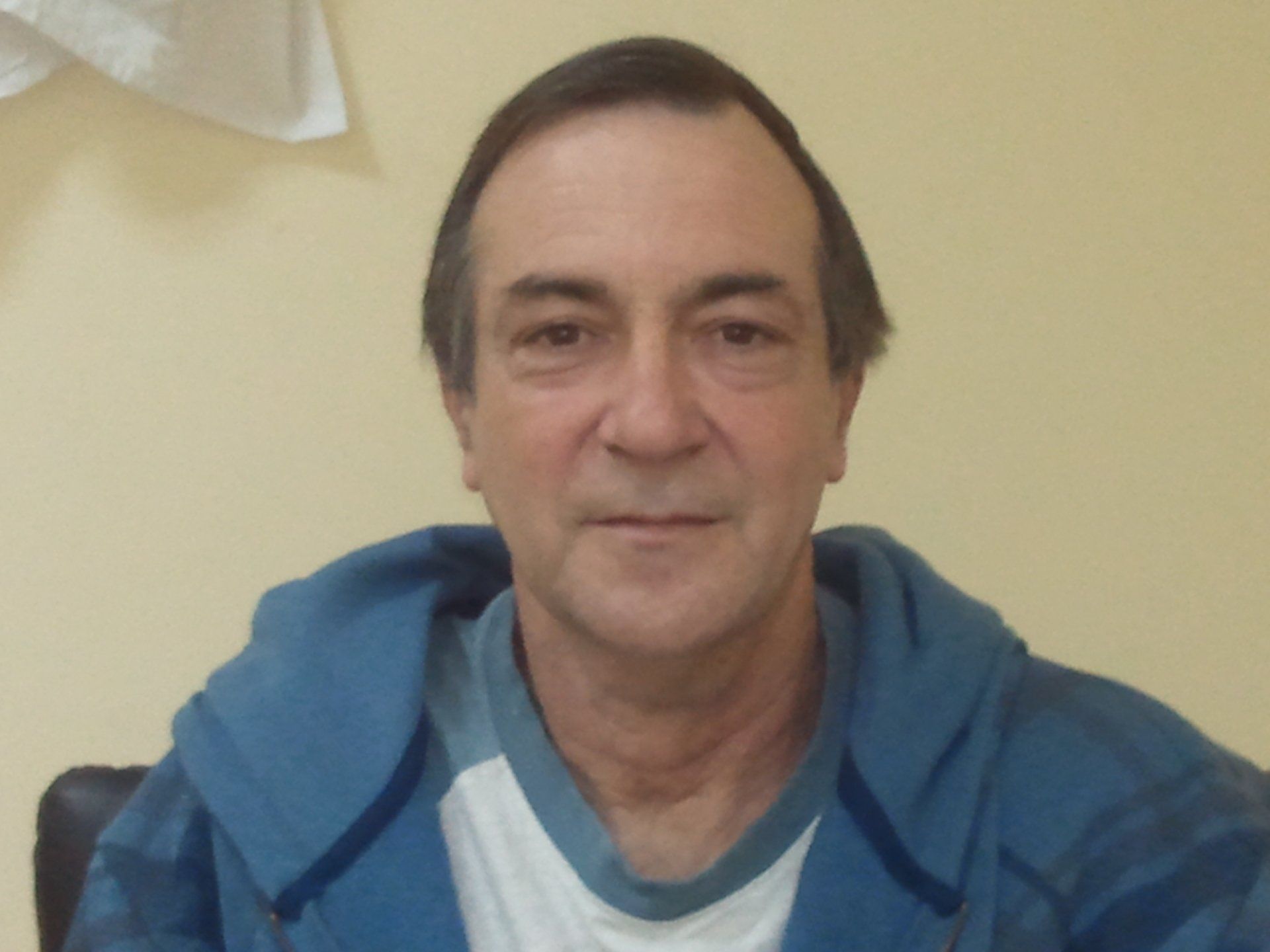 Man in a blue hoodie sitting indoors against a beige wall, looking at the camera.