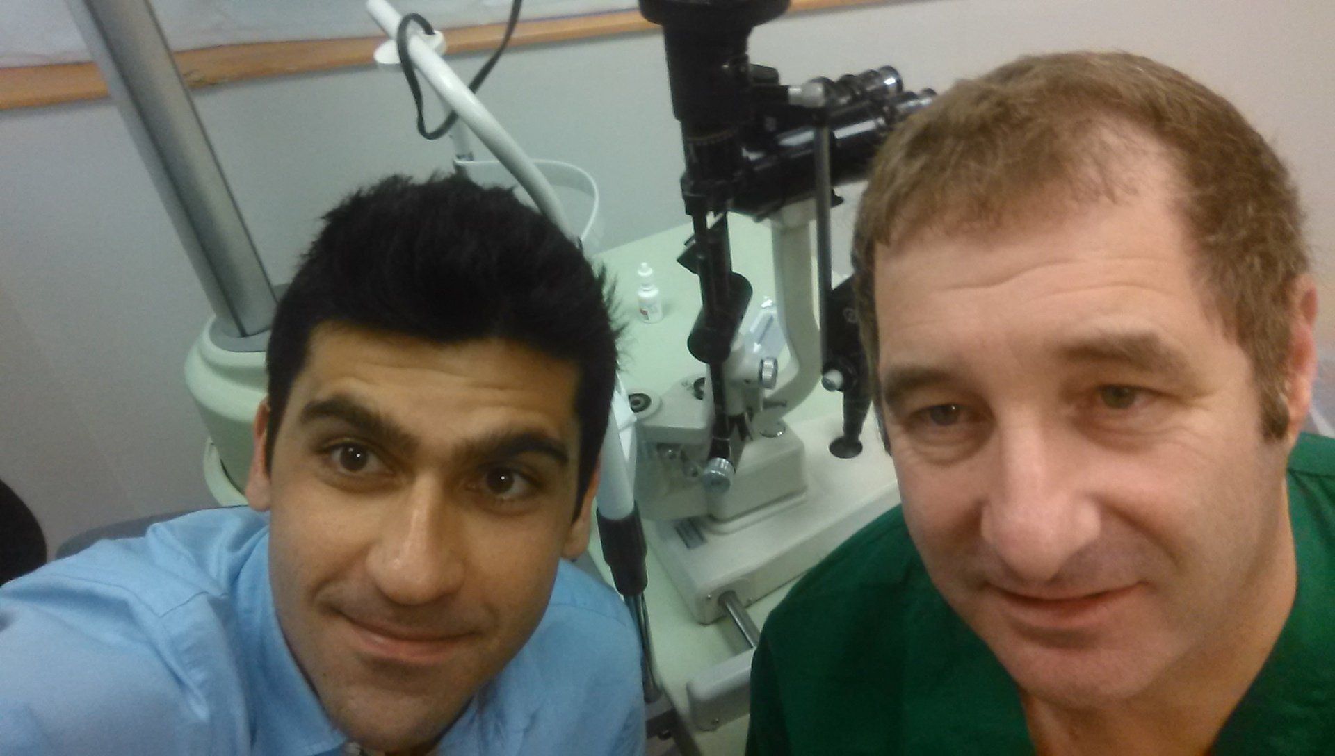 Two people smiling in a medical exam room, with an ophthalmic device behind them.