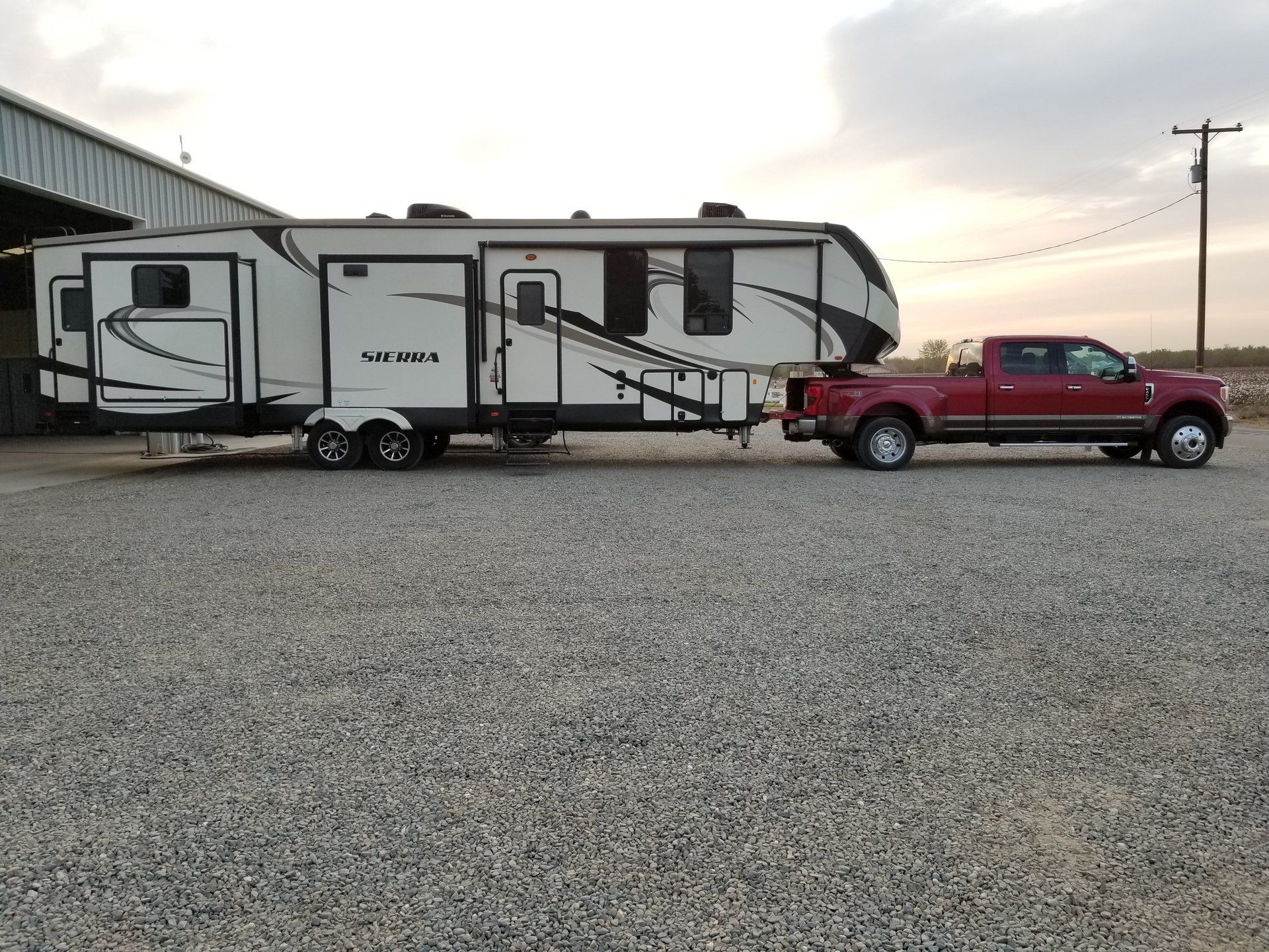 A red truck is towing a trailer in a gravel lot.
