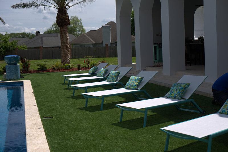 Poolside lounge chairs on artificial turf, beneath a white arched structure, next to a pool.