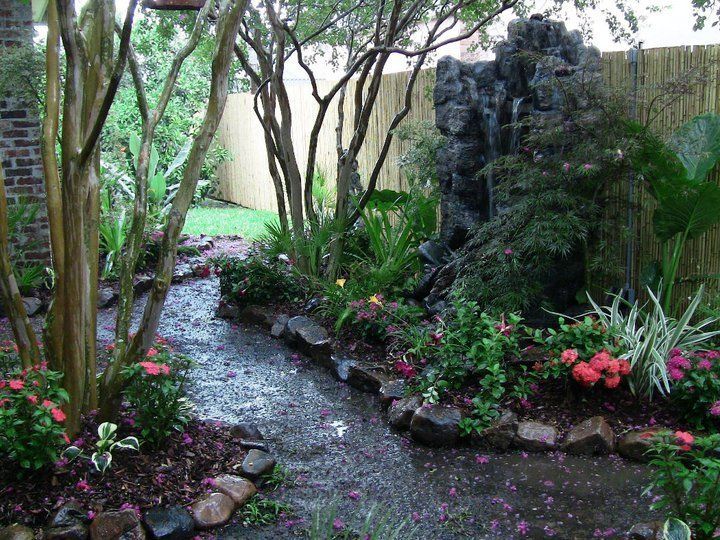 Pathway through lush garden with waterfall feature, bordered by trees and flowers.