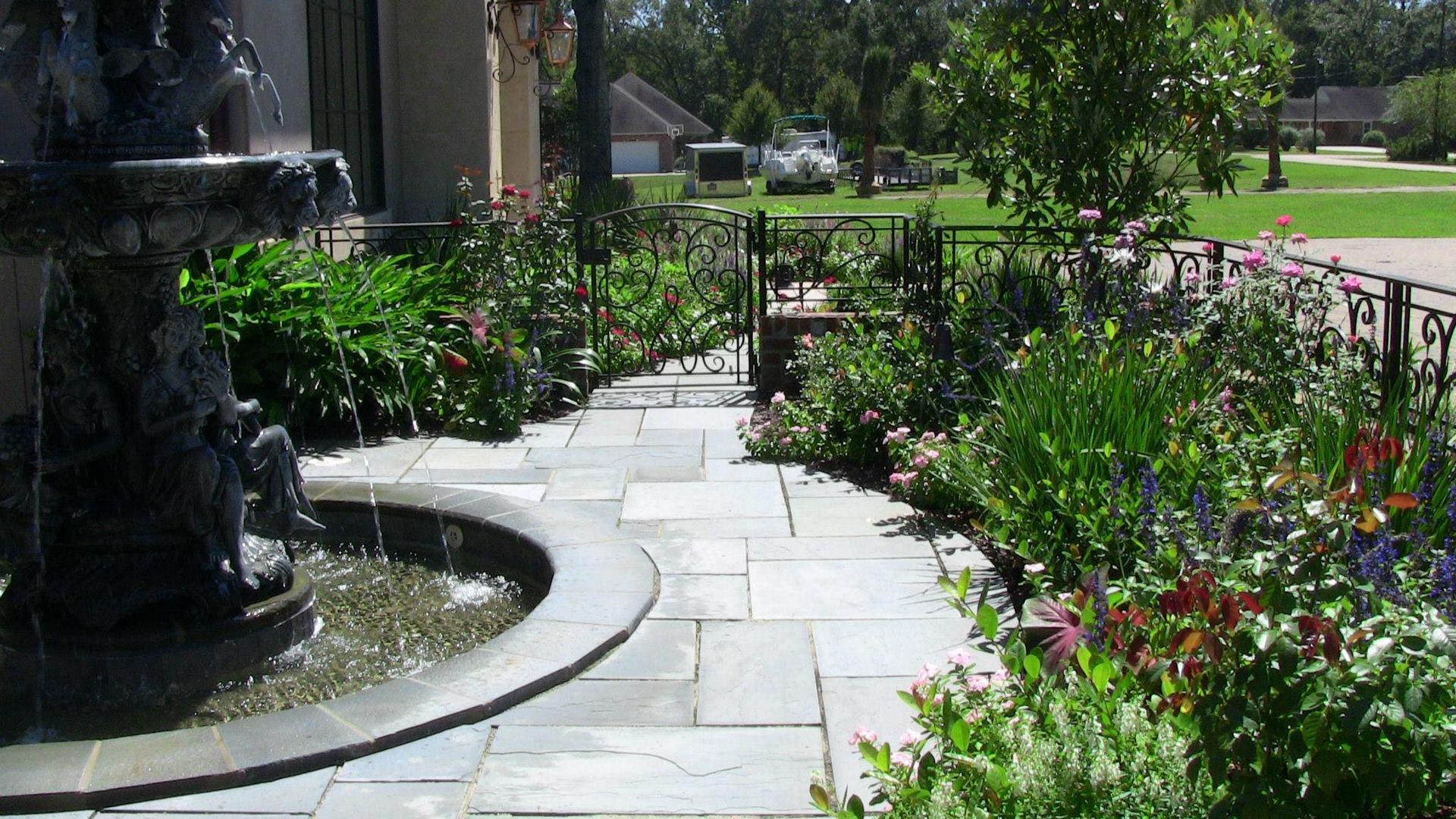 Stone path lined with flowers leads from a fountain to a wrought iron fence and a grassy field.