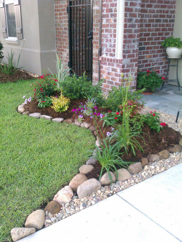 Lush flower bed with varied plants, framed by rocks, next to green lawn and brick column by a building.