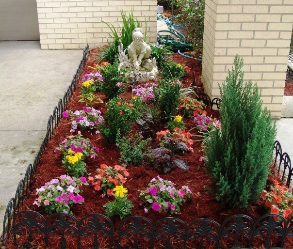 Flower bed with black metal edging, filled with colorful flowers and shrubs, a statue, and brick columns.