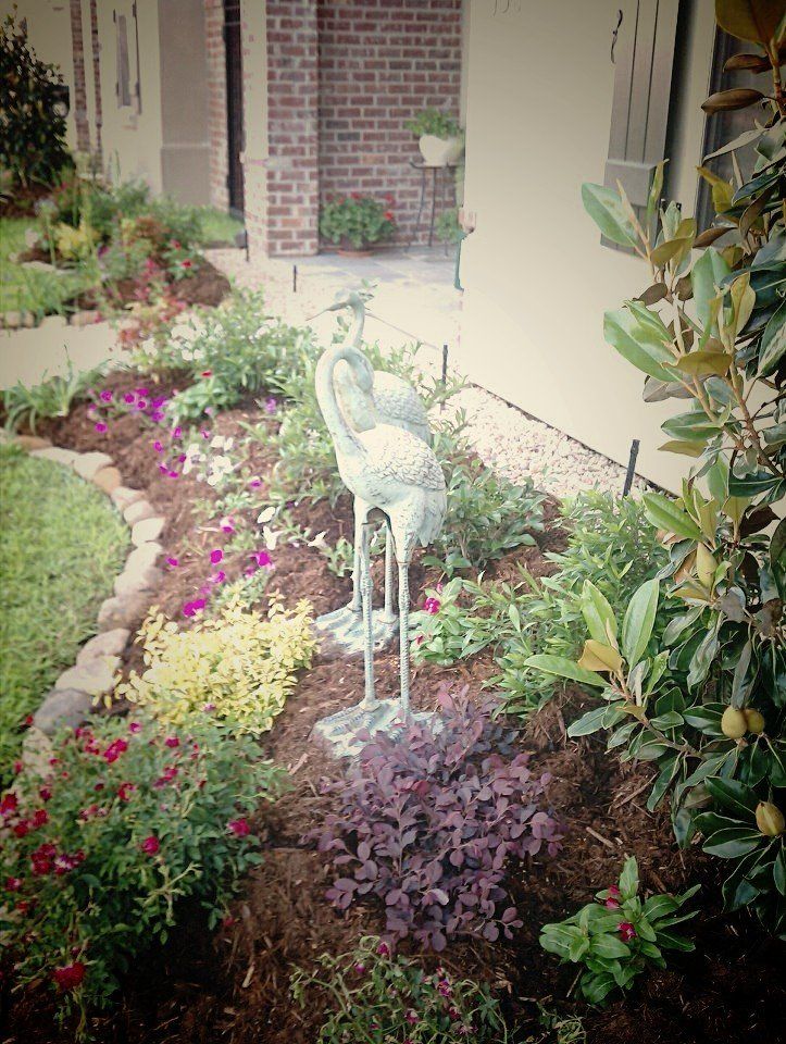 Bronze bird statue in a flower garden with colorful blooms and fresh mulch. Brick wall in the background.