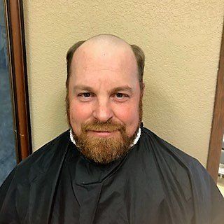 Smiling bald man with a beard wearing a black barber cape against a beige wall