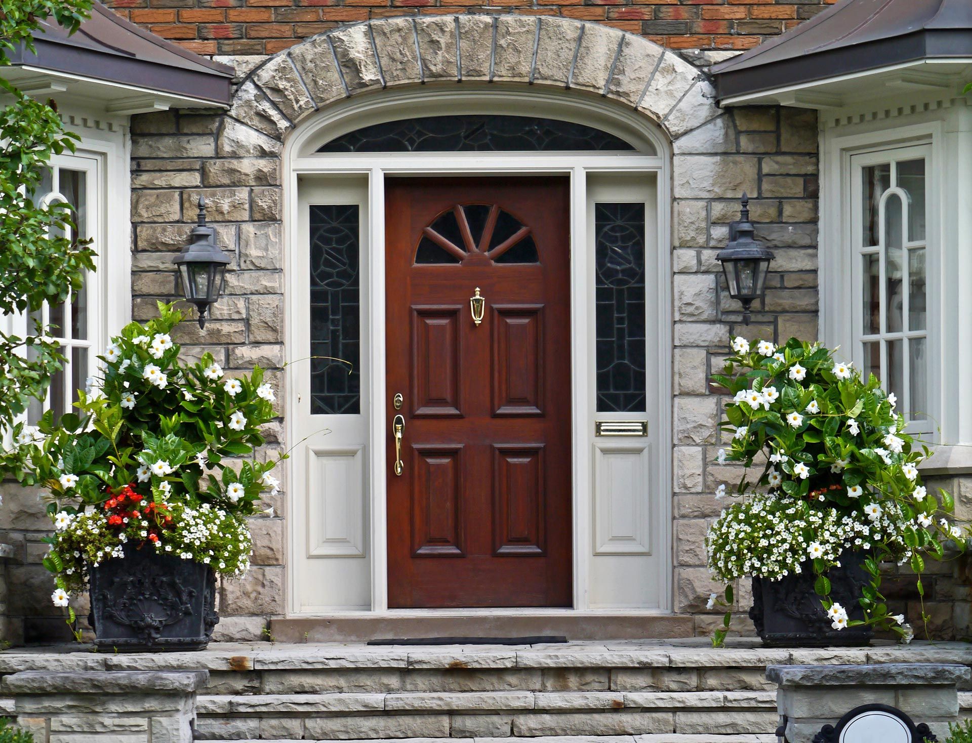 Stone house entrance with a dark wooden door, arched transom window, and flower planters on both sides