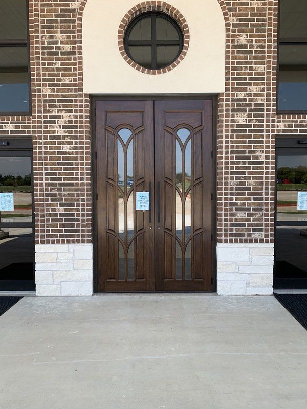 Brick building entrance with dark wooden double doors and a round window above, viewed from a concrete walkway